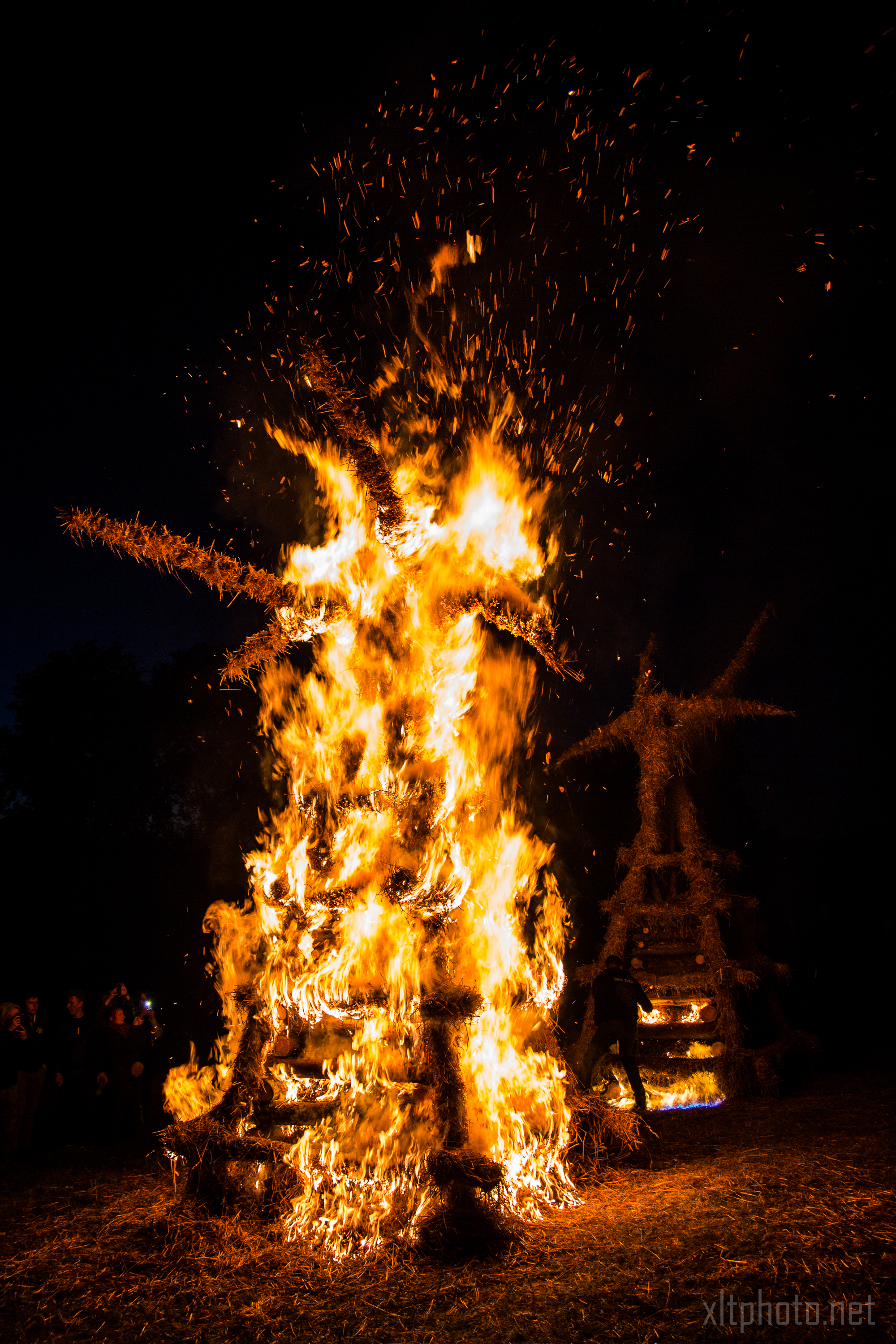 During the 2015 Kilkim Žaibu festival, a large straw effigy is engulfed in bright, intense flames at nighttime, sending sparks and embers into the dark sky. A crowd of spectators watches the bonfire from a distance. Another smaller burning effigy is visible in the background. The ground is covered in straw.