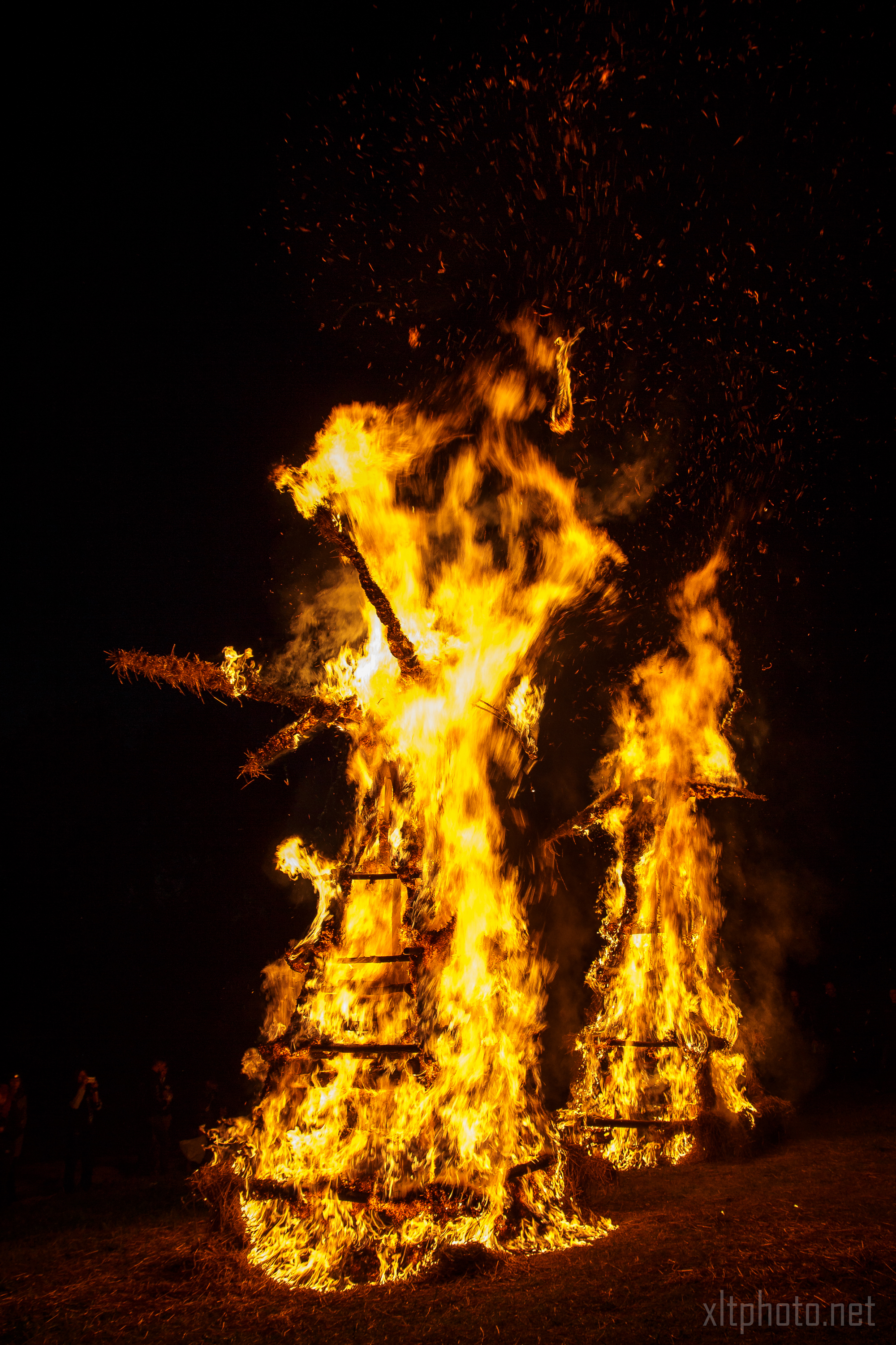 Two large wooden structures engulfed in bright, intense flames during the night at the 2015 Kilkim Žaibu music festival. The fire illuminates the dark surroundings, casting a dramatic glow. A small crowd of silhouetted figures can be seen in the distance observing the burning structures.