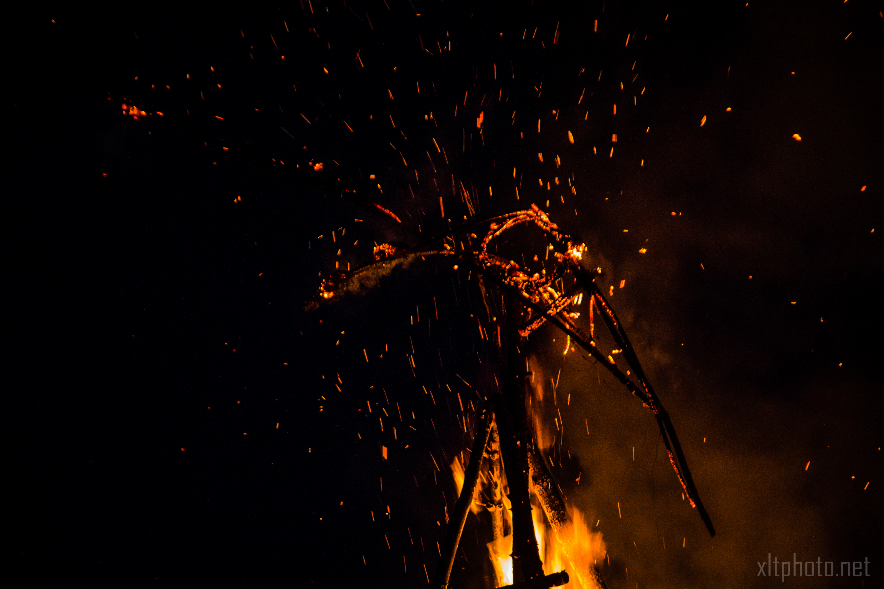 A large fire burns in the dark, with bright orange and yellow flames illuminating a skeletal structure within the blaze. Sparks fly upward, creating a dramatic and intense scene. The image captures the dynamic and destructive nature of the fire against a pitch-black background at Kilkim Žaibu 2015 music festival.