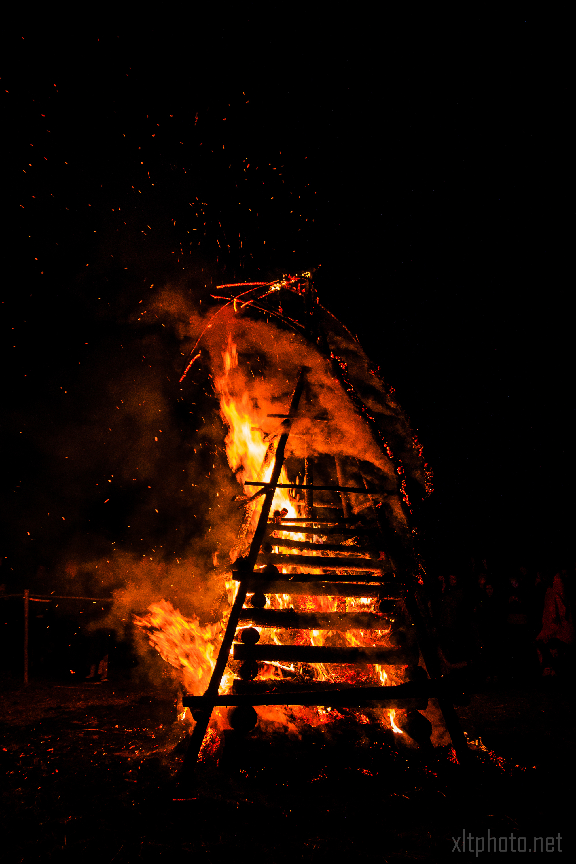A large wooden structure burns intensely in the night at the 2015 Kilkim Žaibu music festival, emitting bright flames and sparks. The dark sky and surrounding area are illuminated by the fire, with silhouettes of people visible in the background. The structure appears to be a tall, slatted frame built from logs and branches.