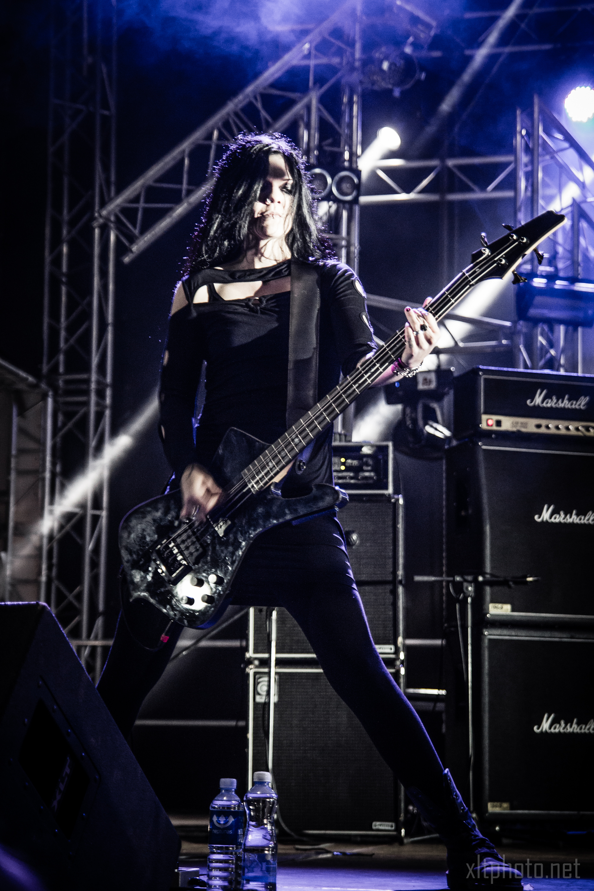 A musician with long black hair, wearing a black outfit, plays a bass guitar passionately on stage at the 2015 Kilkim Žaibu festival. Surrounded by metal scaffolding and numerous Marshall amplifiers, the stage is lit with strong white and blue lights, while water bottles lie on the floor in the foreground.