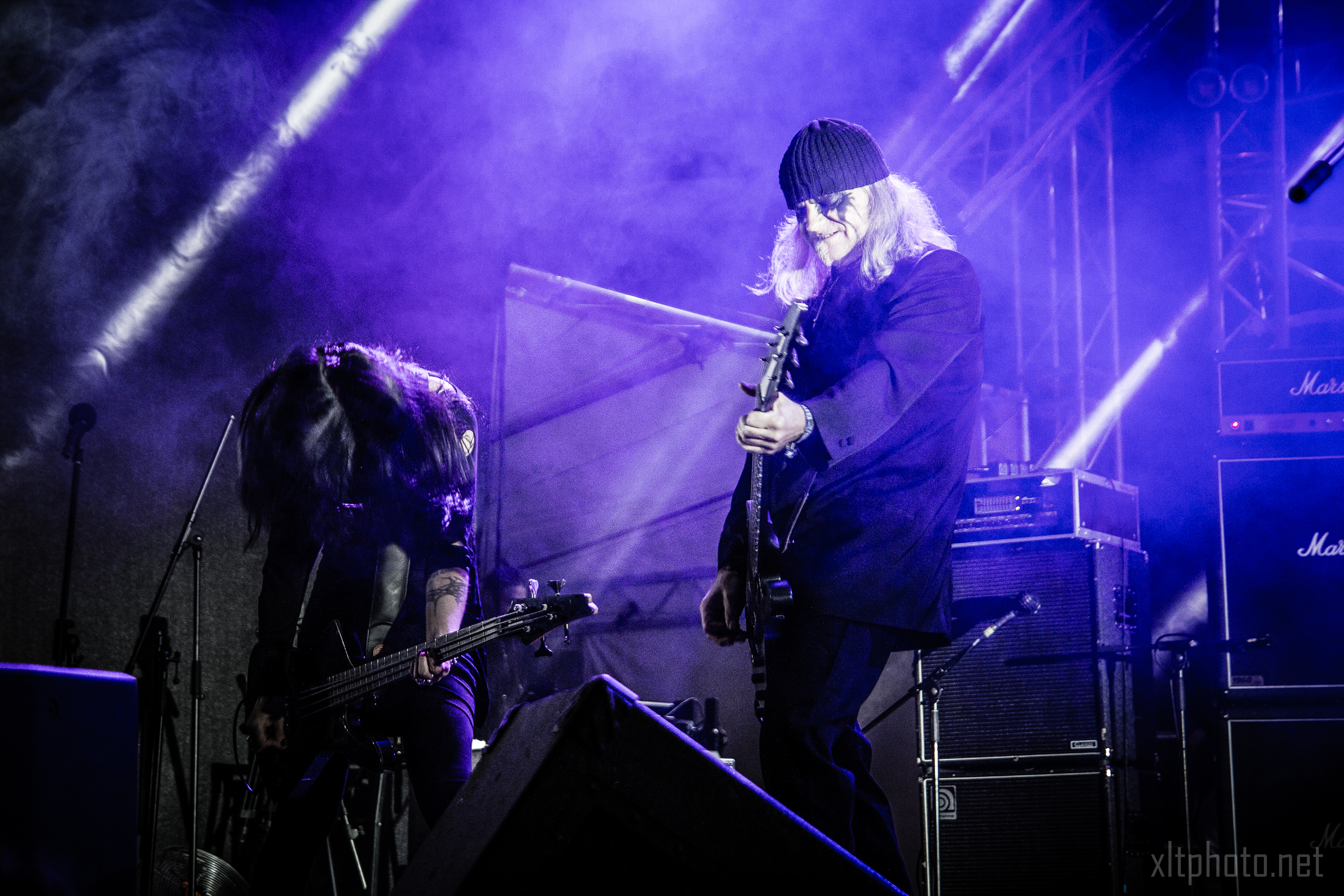 Two musicians perform on a dimly lit stage with blue and purple spotlights at Kilkim Žaibu 2015. The guitarist on the right wears a beanie and dark coat, playing intently, while the bassist on the left, with long hair covering their face, is deeply engrossed in playing. Amplifiers and stage lighting are visible in the background.