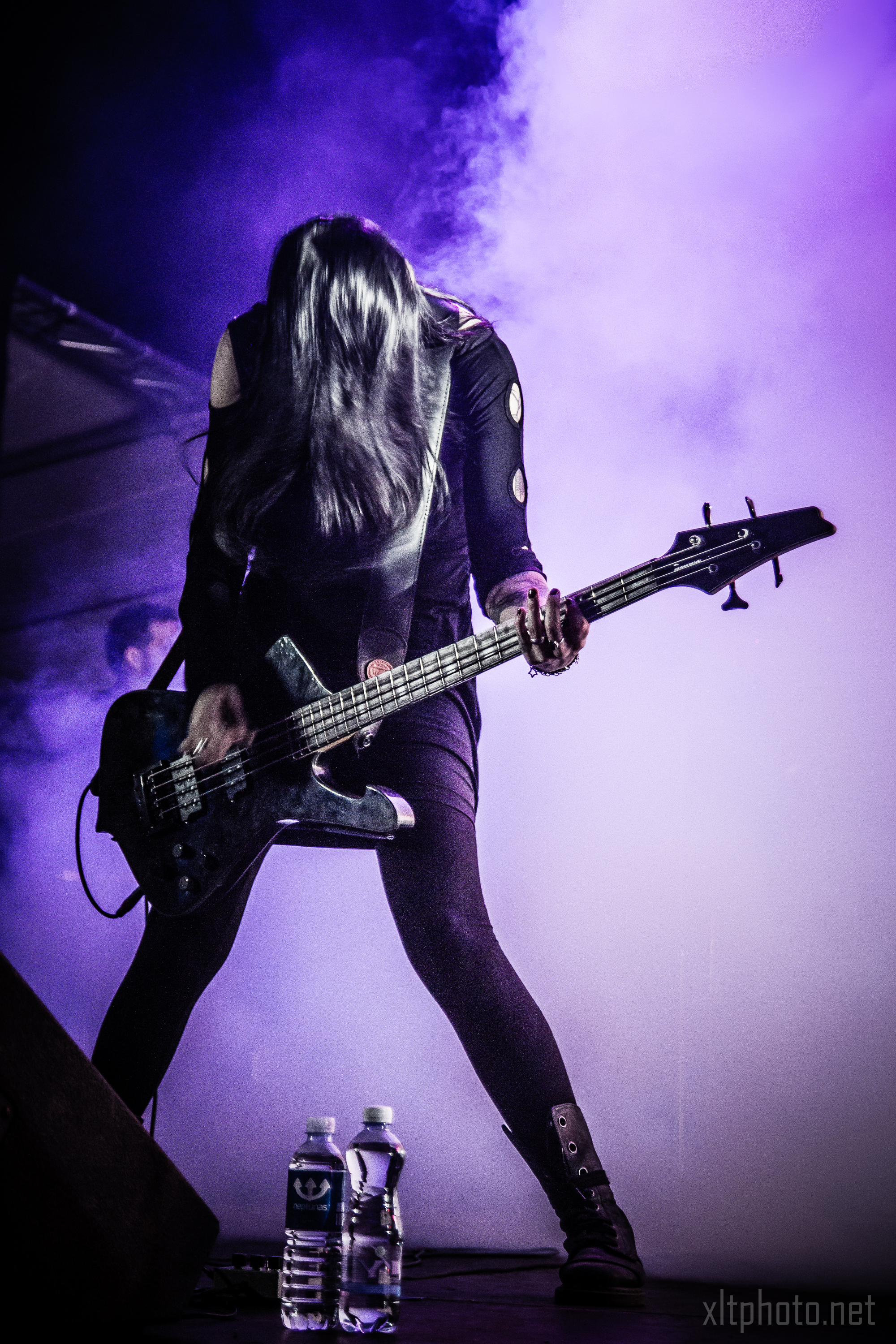 At the 2015 Kilkim Žaibu festival, a woman wearing a black outfit passionately plays a bass guitar on stage, surrounded by a purple mist. Her long hair obscures her face as she leans forward, immersed in her performance. Two water bottles and effects pedals are visible on the ground in front of her. Lighting highlights her dynamic presence.
