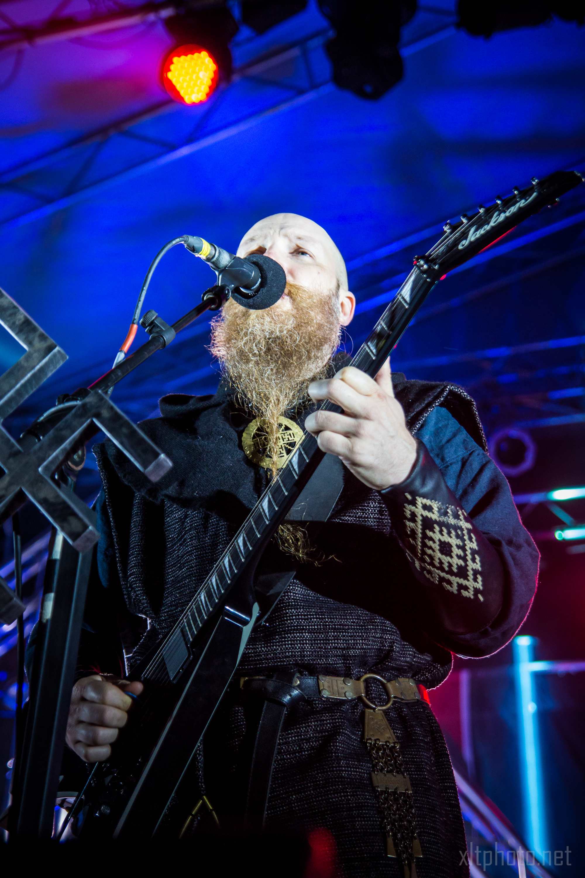 A musician with a long, curly beard plays an electric guitar on stage at the 2015 Kilkim Žaibu festival. He is singing into a microphone and dressed in a dark, medieval-inspired outfit. The background is lit with blue and red stage lights, creating an atmosphere that suggests a live concert performance.