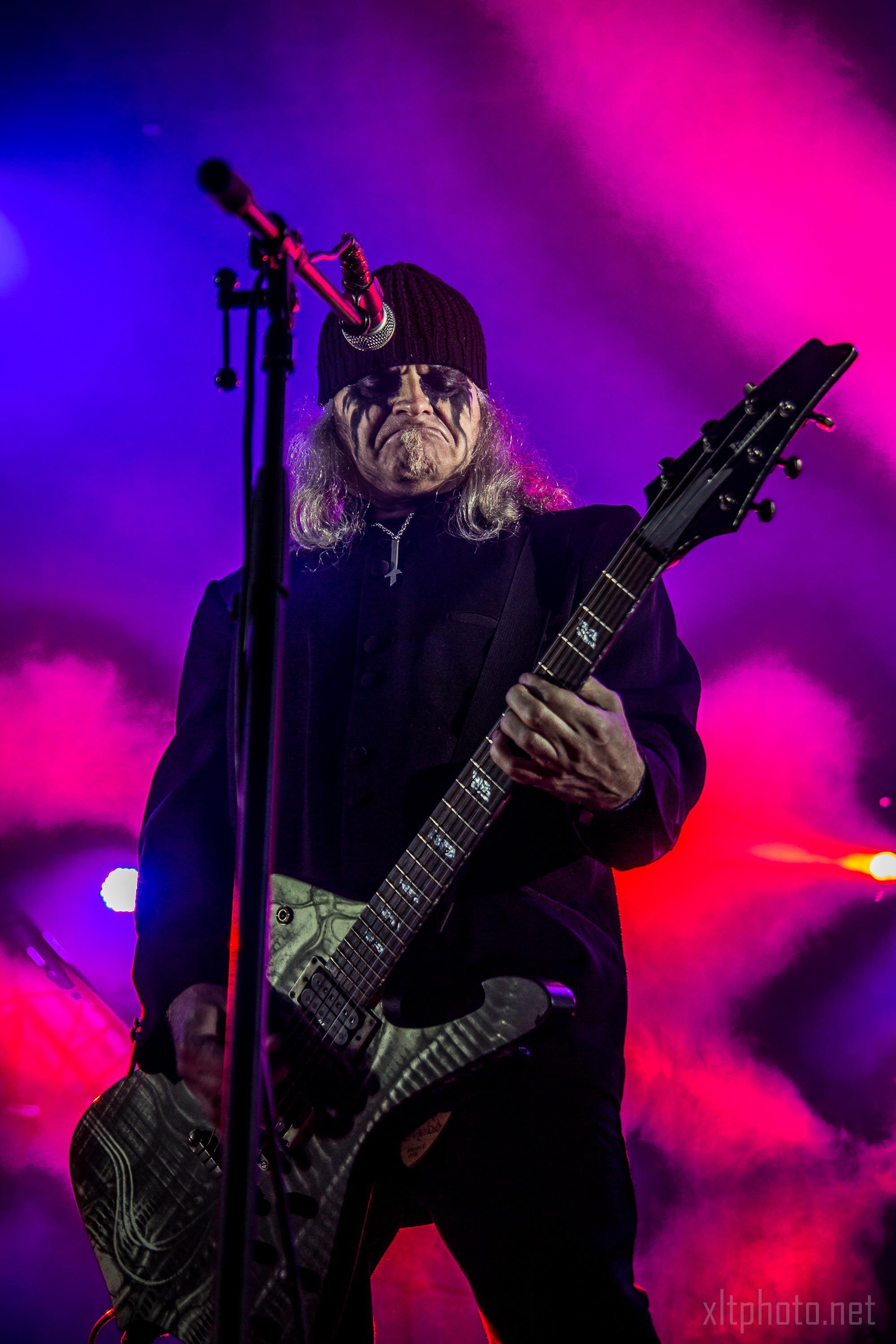 A musician with long hair and face paint is playing a guitar on stage. He wears a dark beanie, sunglasses, and an all-black outfit. Vibrant pink and purple stage lights create a dramatic background, casting a colorful glow and enhancing the intensity reminiscent of Kilkim Žaibu 2015.