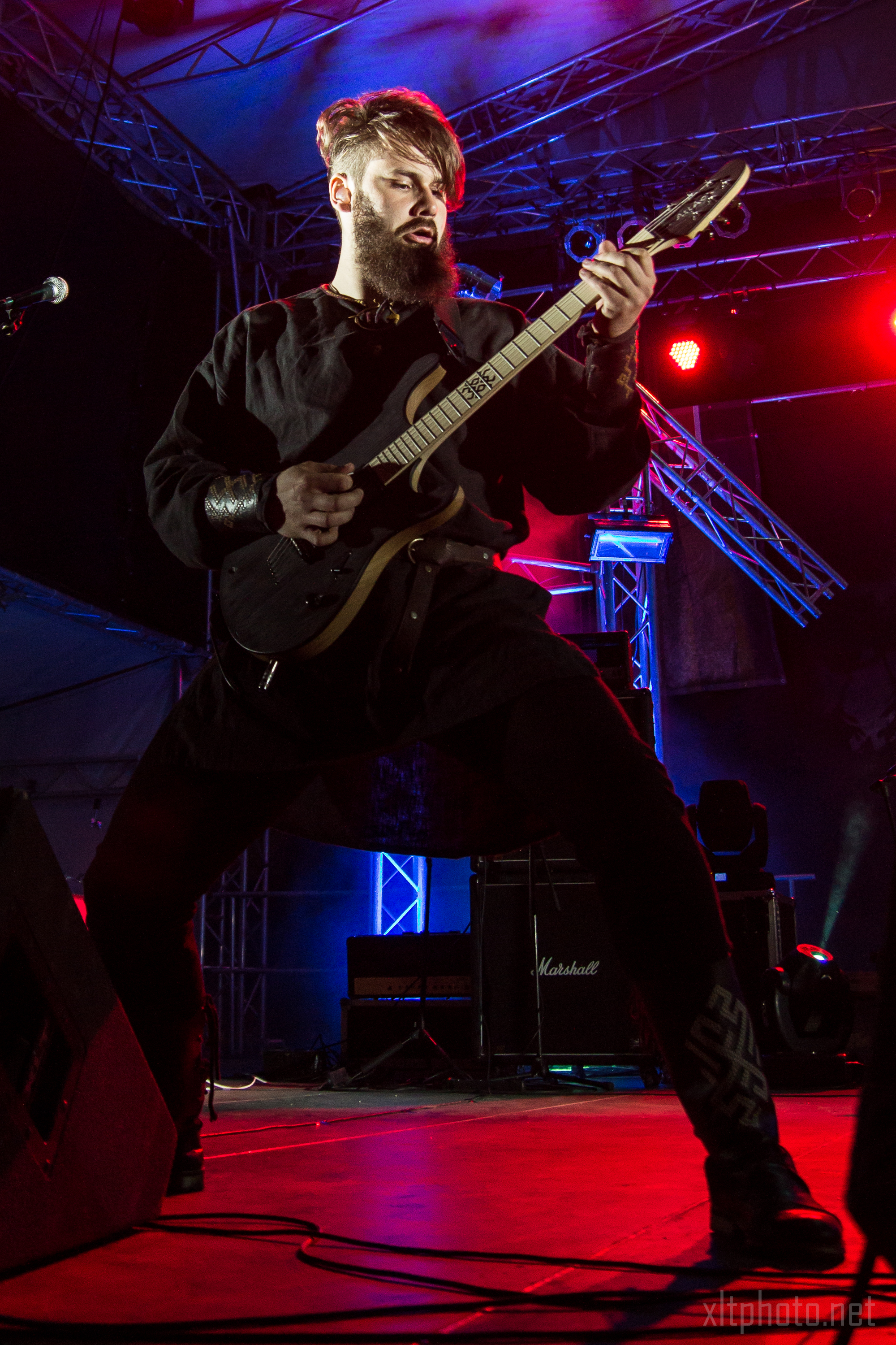 A musician with a beard, wearing a black outfit, passionately plays an electric guitar on a dimly lit stage with vibrant red and purple lights. The stage has metal structures and amplifiers in the background. The musician stands in a wide stance, fully immersed in the performance at Kilkim Žaibu 2015.