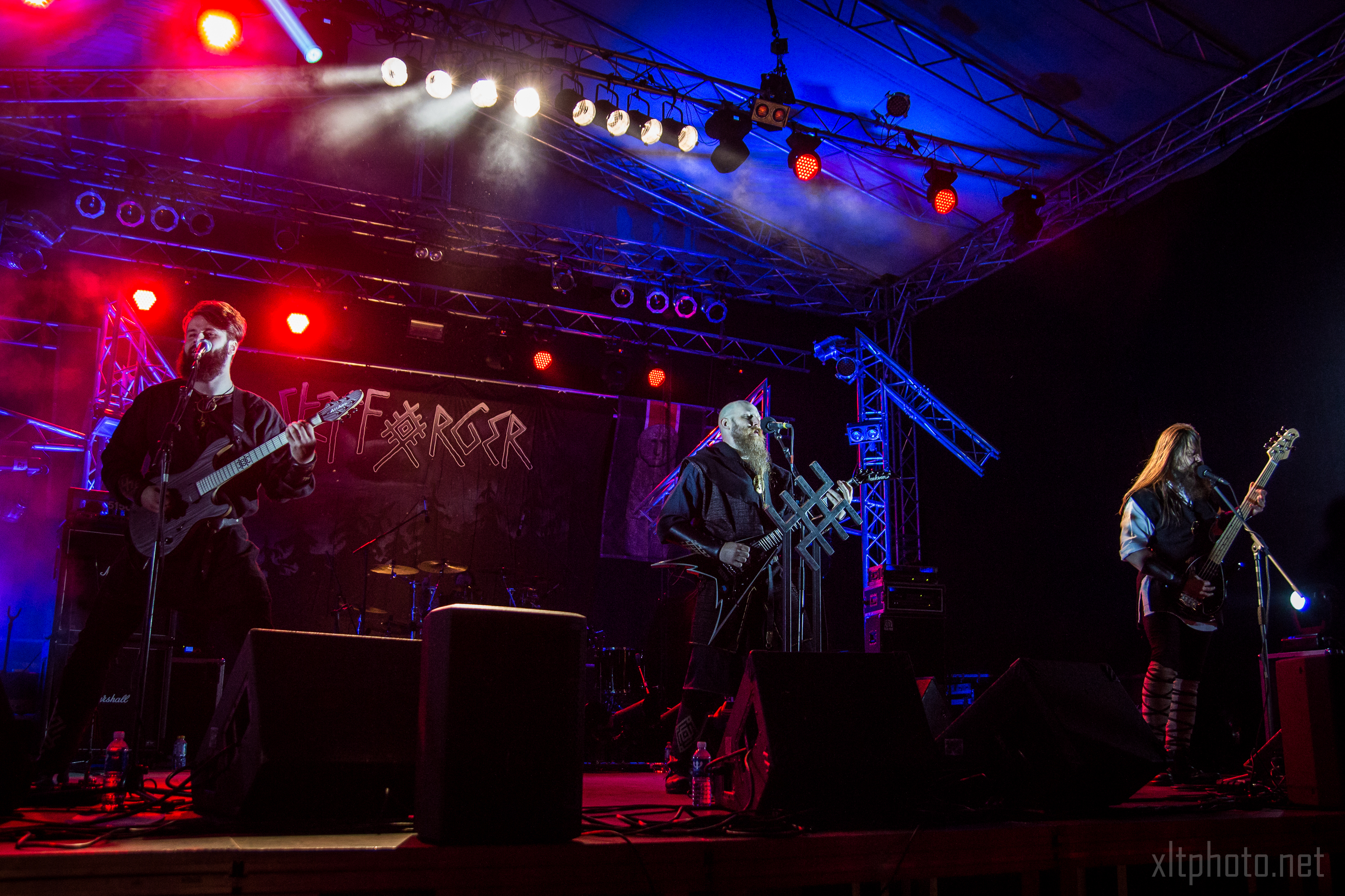 A band performing live on stage at the 2015 festival under colorful lighting. Three members are visible, each playing a string instrument, with the middle one singing into a microphone. The stage is equipped with various lighting and sound equipment, and the backdrop proudly displays "Kilkim Žaibu.