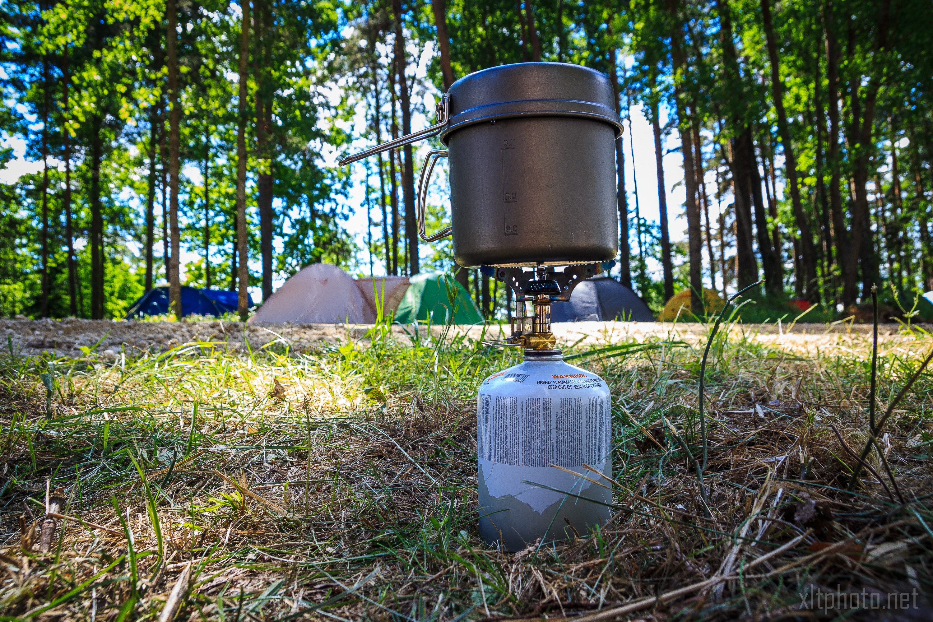 A portable camping stove with a small fuel canister heats a pot with a ladle inside. The stove is set on a grassy and wooded area with several colorful tents visible in the background, reminiscent of a lively 2015 Kilkim Žaibu festival. The scene is bright and sunny, with tall trees providing a green canopy.