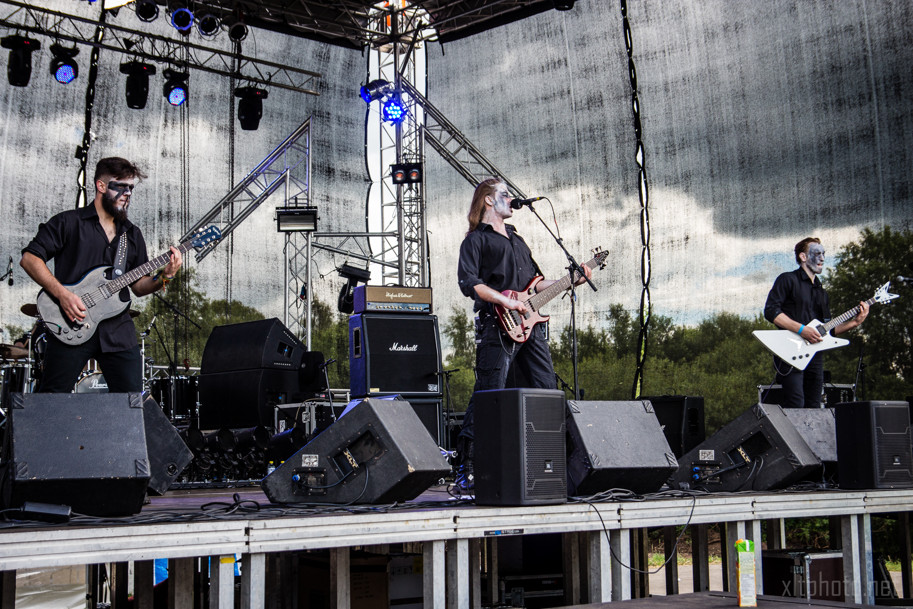 At Kilkim Žaibu festival in 2015, four musicians dressed in black, with painted faces, perform on an outdoor stage. Two play electric guitars, one plays bass, and the other plays drums. The stage features a metal frame, lighting equipment, and speakers. Trees are visible in the background. The sky is overcast.