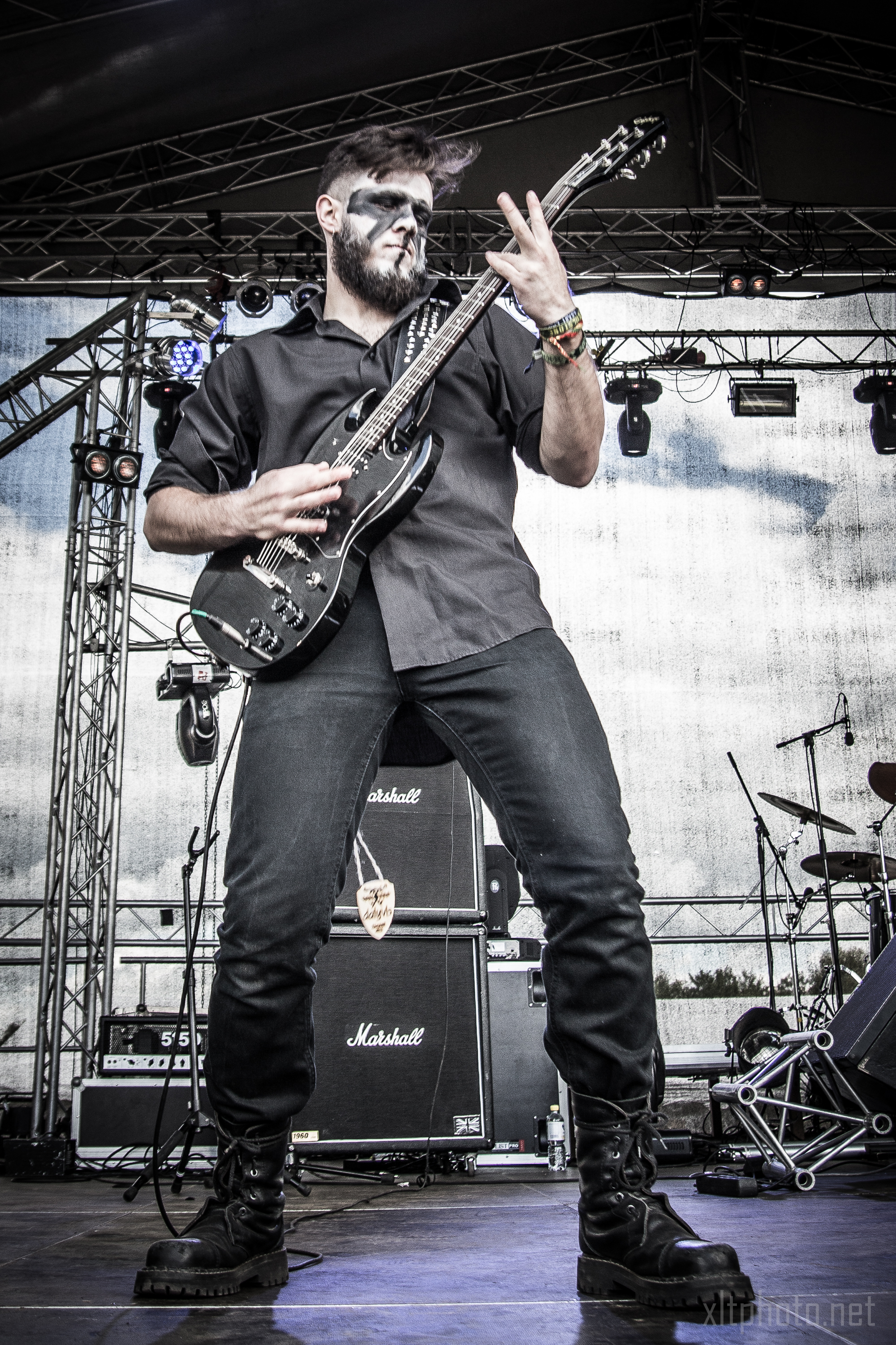 A guitarist with face paint performs passionately on stage at the 2015 Kilkim Žaibu festival. He wears black clothing and combat boots, playing an electric guitar. The background features large Marshall amplifiers and stage lighting equipment against a partially cloudy sky.