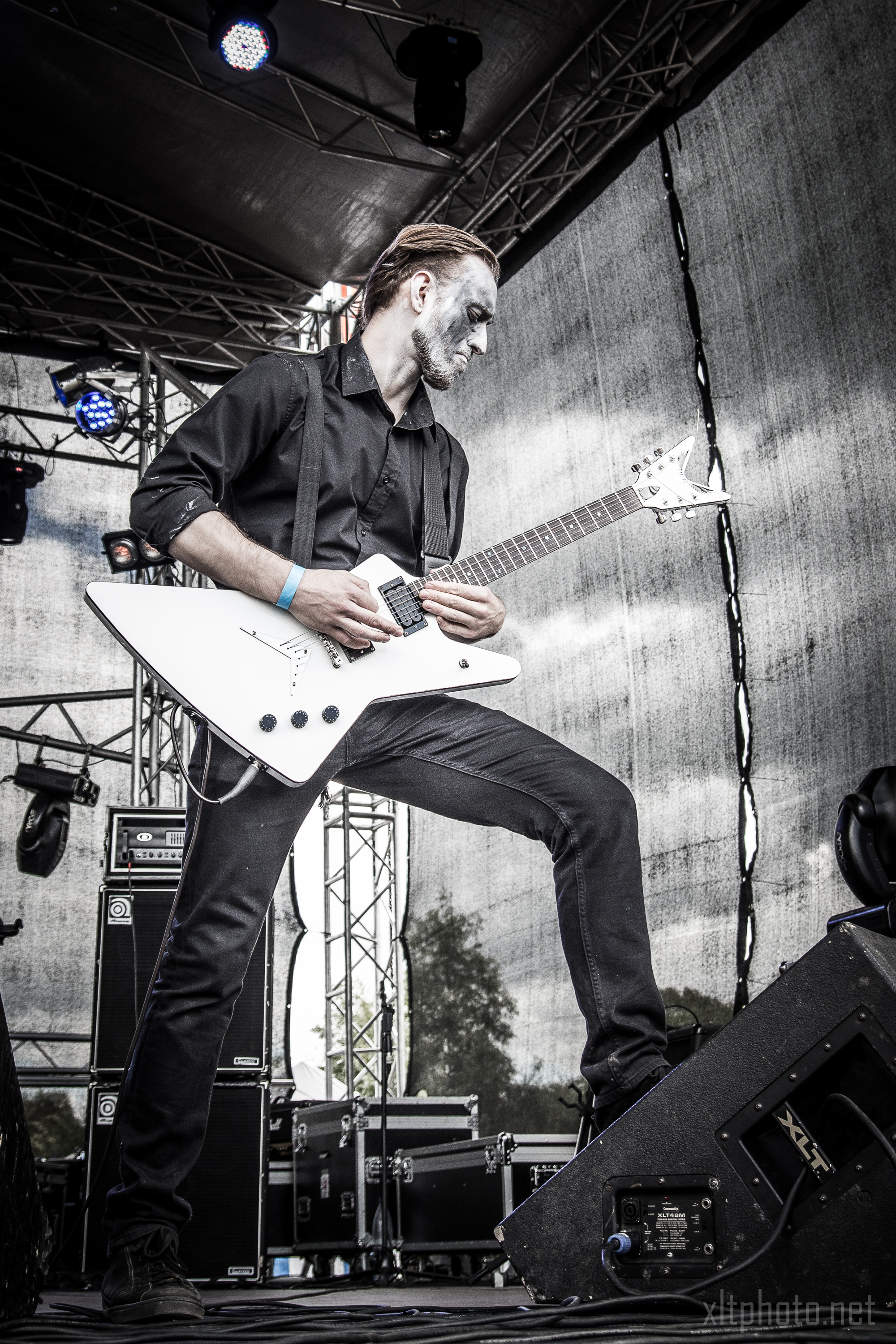 A musician with white face paint is playing a white electric guitar on an outdoor stage at the 2015 Kilkim Žaibu festival. He's dressed in black, including a black shirt and pants, and stands in a powerful stance with one foot on an amplifier. The background includes stage equipment and a mesh backdrop filtering daylight.