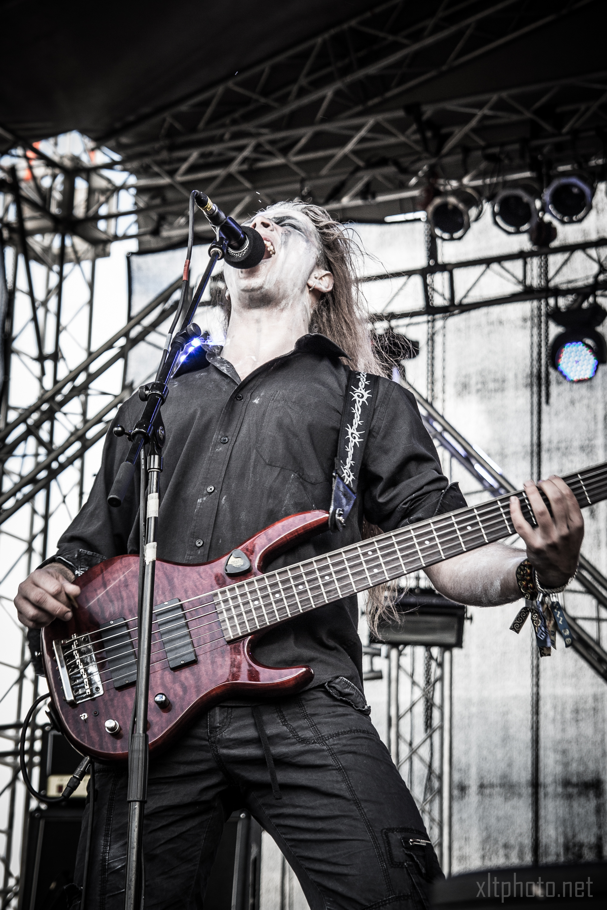 At the 2015 Kilkim Žaibu festival, a musician with long hair passionately sings into a microphone while playing a six-string bass guitar on an outdoor stage. He is wearing a black shirt and has several wristbands on his right wrist, framed by the stage's scaffolds and lighting equipment in the background.
