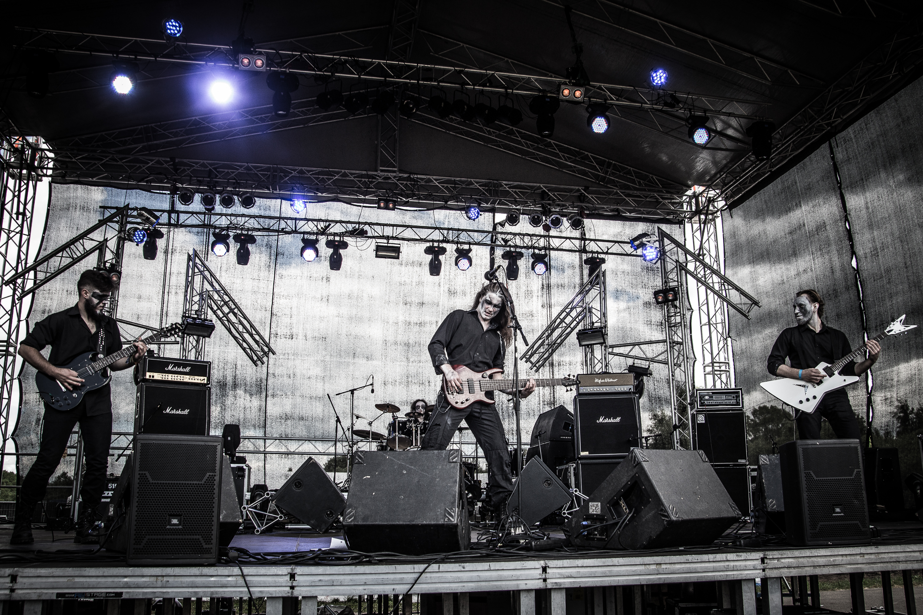 A rock band performs energetically on an outdoor stage at the 2015 Kilkim Žaibu, surrounded by musical equipment and illuminated by blue and white stage lights. Four musicians play electric guitars and bass, with a drum set visible in the background. They are under a large, metal-framed canopy.