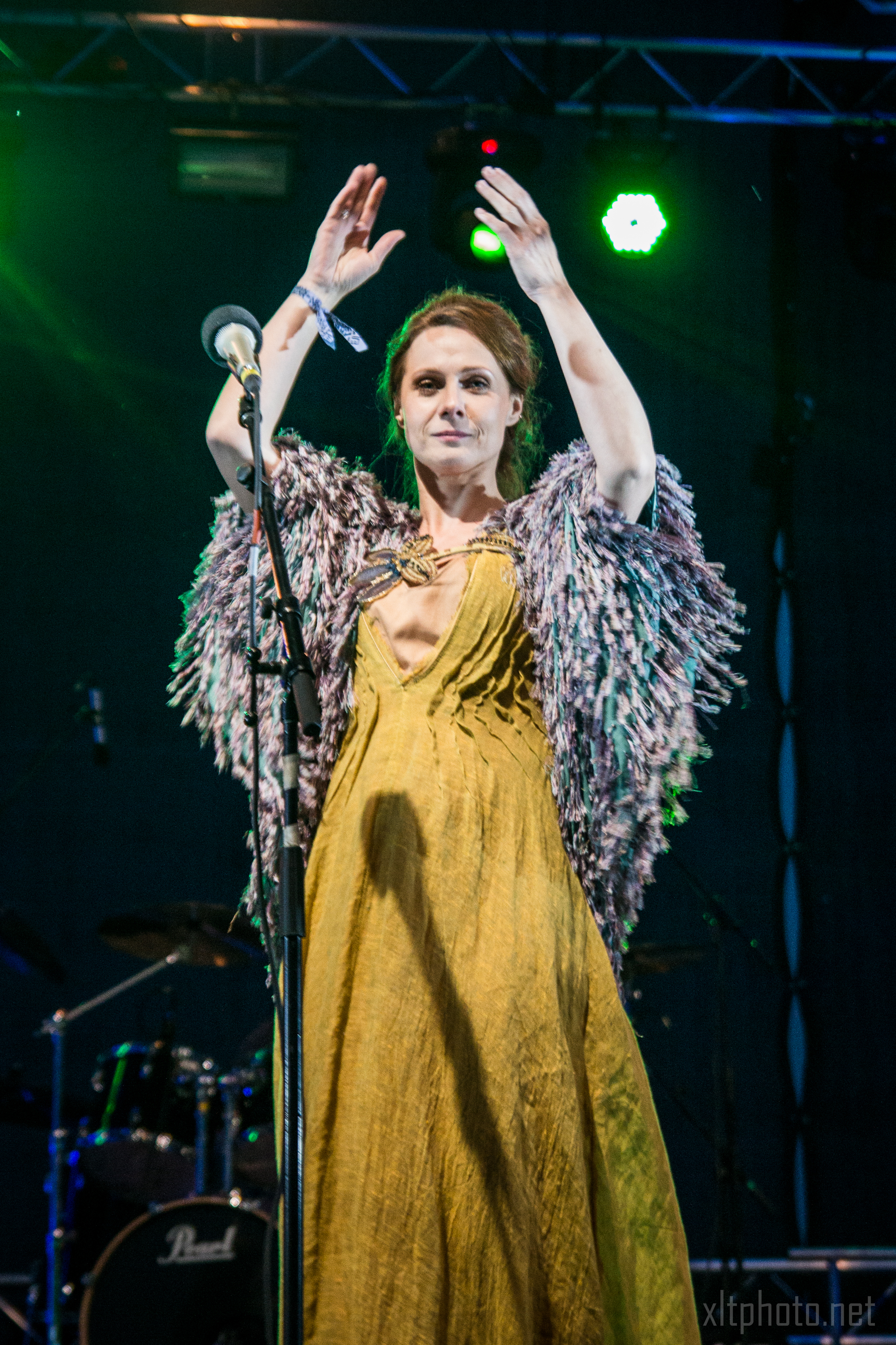 A woman stands on stage illuminated by green stage lights at the 2015 Kilkim Žaibu festival. She wears a flowing, mustard yellow dress and a shawl with feathery textures. Her arms are raised, and she stands in front of a microphone. The background shows part of a drum set, and the atmosphere suggests a live music performance.