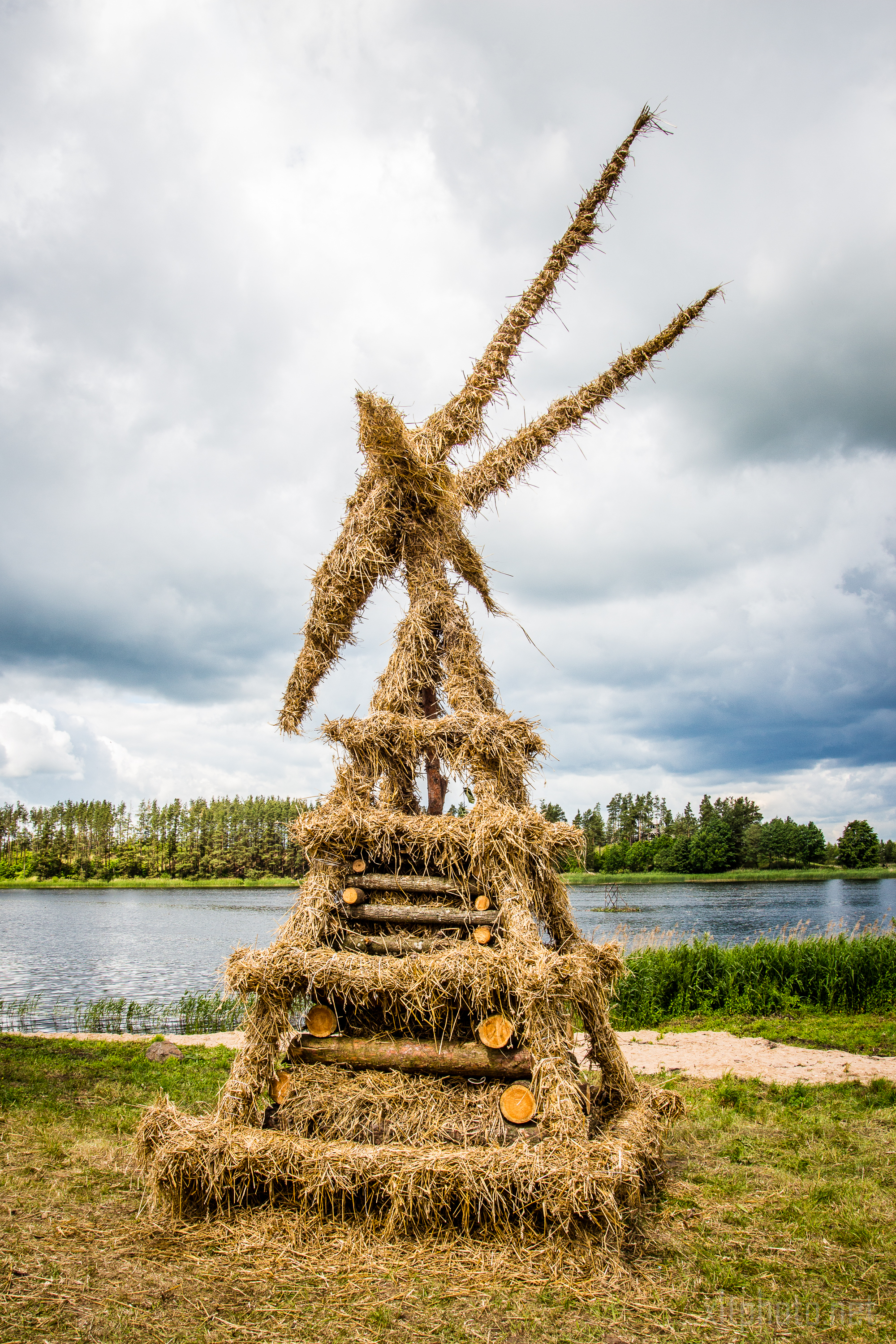 A large, artistic sculpture made of hay and logs stands on grassy ground near a lake. The structure is pyramid-shaped with triangular extensions at the top, resembling a figure holding spears. Created in 2015 for the Kilkim Žaibu festival, it sits under an overcast sky with trees lining the opposite bank of the lake.