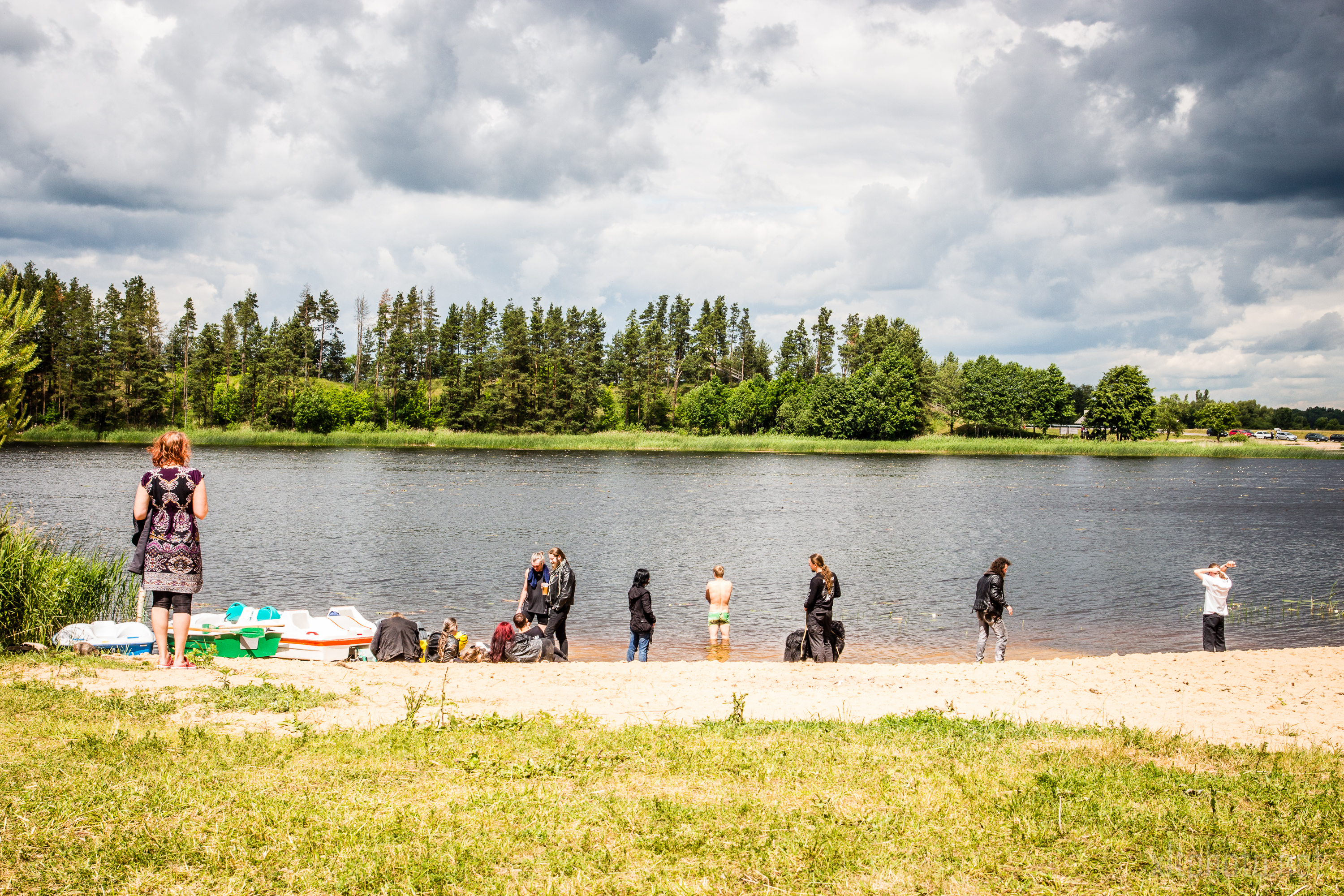 A group of people stands on a sandy beach beside a calm lake, with lush green trees lining the opposite shore. Some individuals are near the water while others are on the sand. Several paddle boats are on the shore, and the sky above is partly cloudy with patches of blue, reminiscent of Kilkim Žaibu 2015.