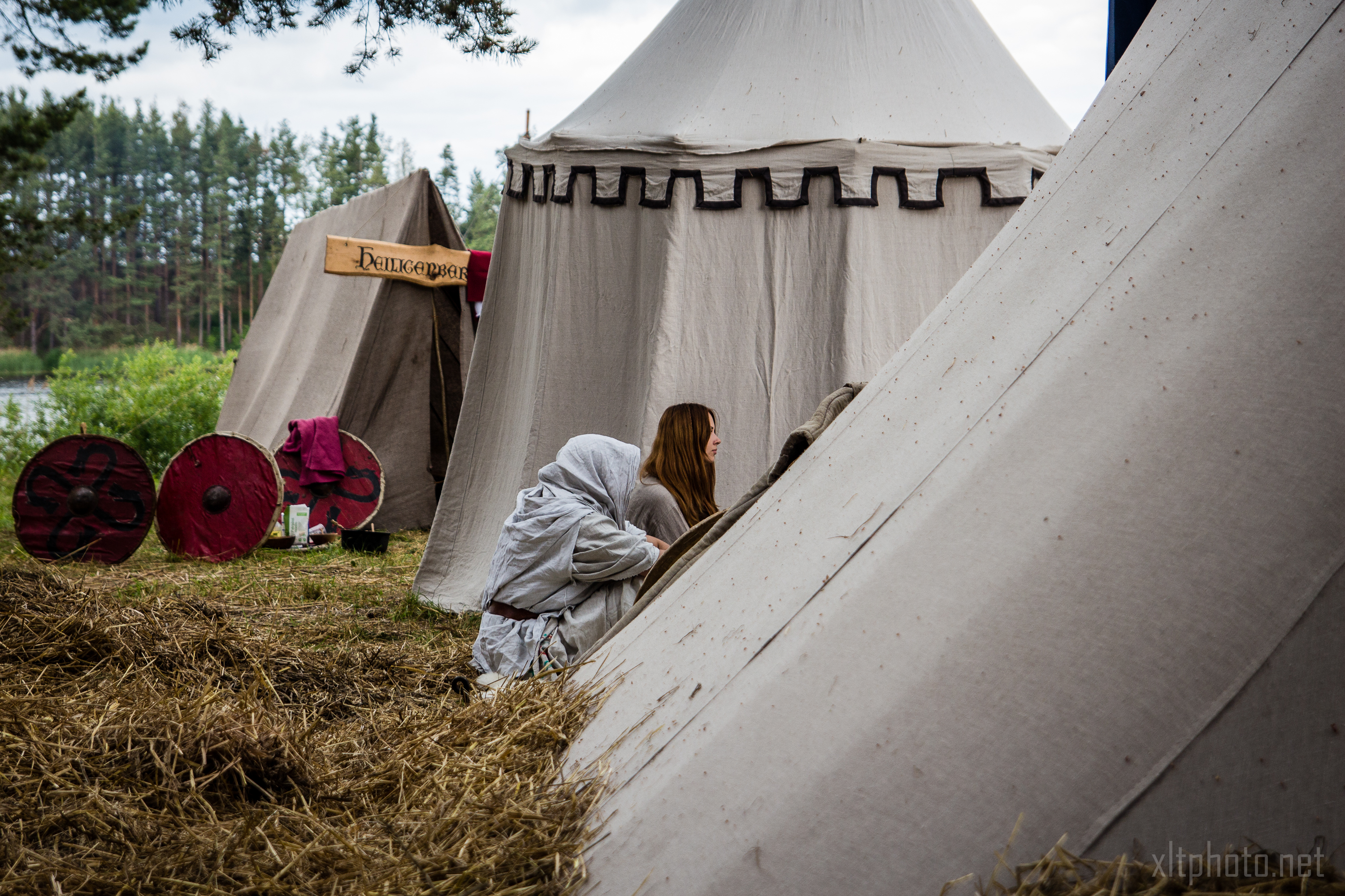 People in medieval-style clothing sit and interact near large, beige canvas tents at an outdoor camp setup during Kilkim Žaibu 2015. Two red wooden shields lean against one tent in the background, and a wooden sign with illegible text hangs nearby. The area is surrounded by trees and covered with straw.