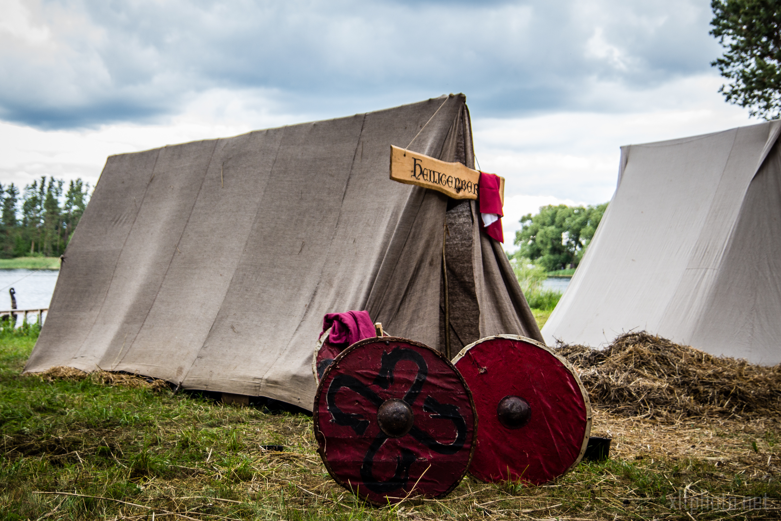 A medieval-themed camp scene from Kilkim Žaibu 2015 features a canvas tent with a wooden sign above its entrance. Two painted wooden shields, one red with a black pattern, lean against the tent. Another similar tent and a pile of hay are in the background, with a river and trees visible under a cloudy sky.