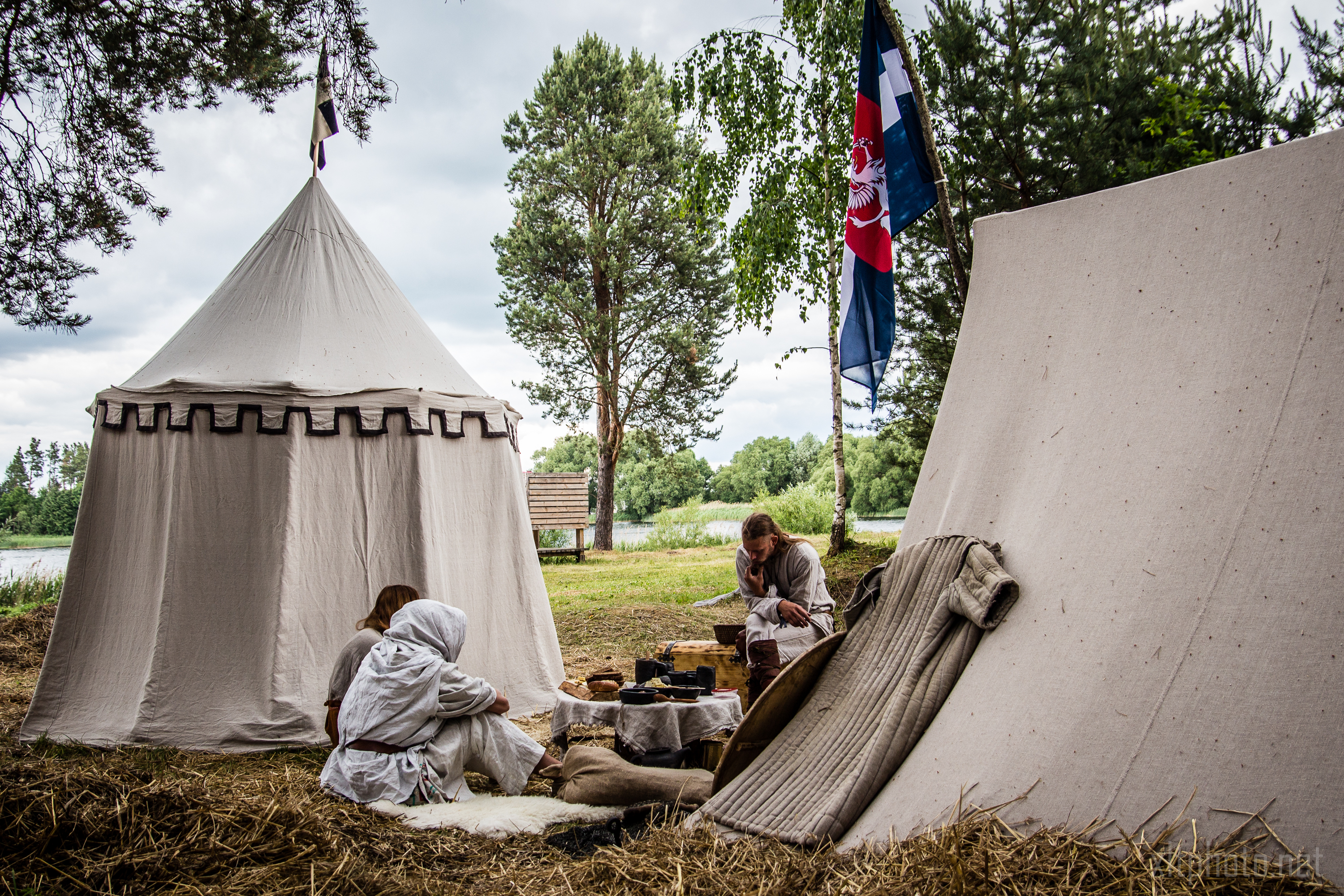 A scene at the 2015 Kilkim Žaibu outdoor historical reenactment camp shows two canvas tents set up among trees. Two people in period clothing sit near a table with wooden items and food. A river, flags, and more trees are in the background. The atmosphere is rustic and tranquil.