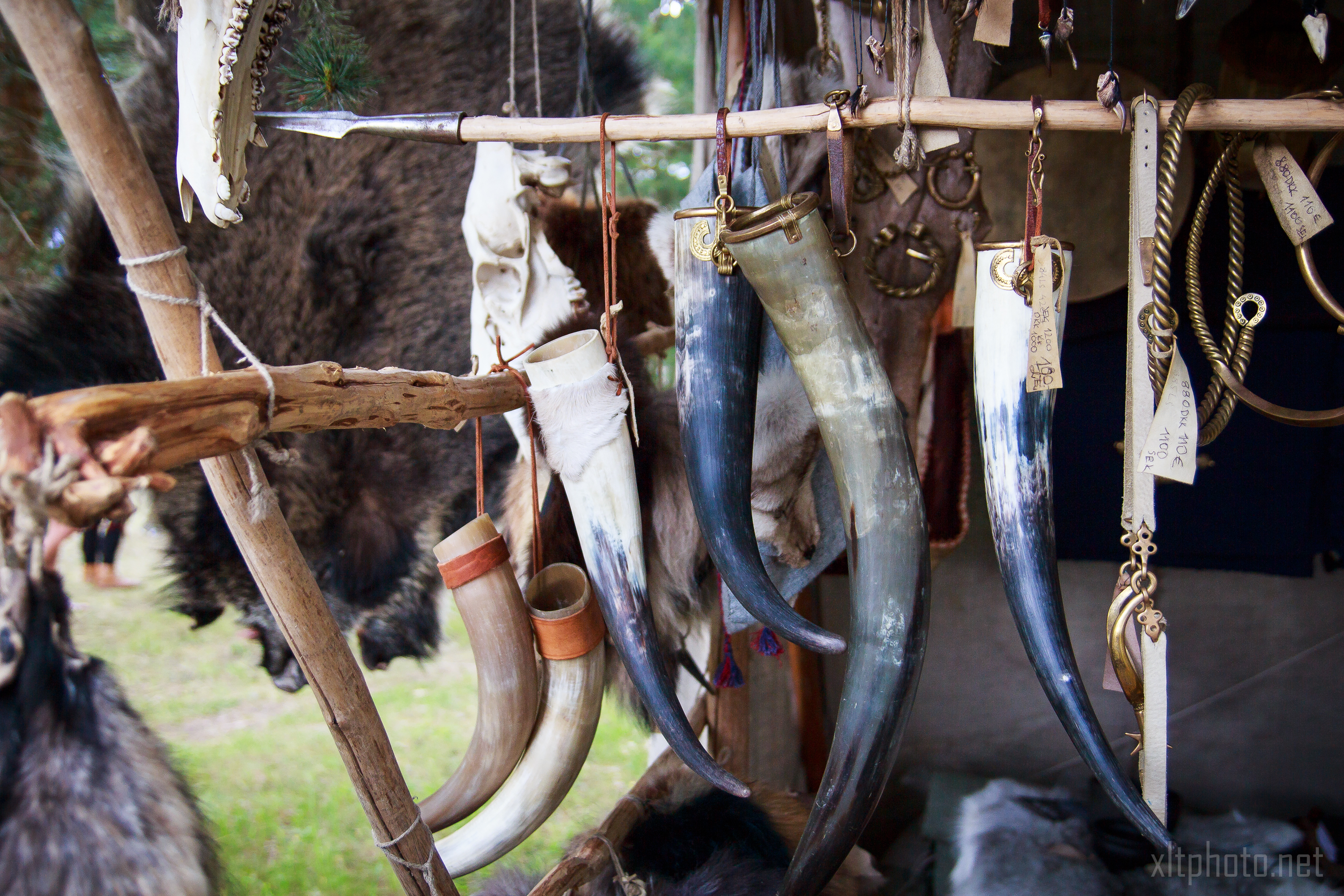 A display of assorted animal horns hangs from a wooden structure at an outdoor market, reminiscent of Kilkim Žaibu 2015. The horns vary in size and color, adorned with leather straps and metal rings. In the background, fur pelts and other traditional items create a rustic, historical ambiance.