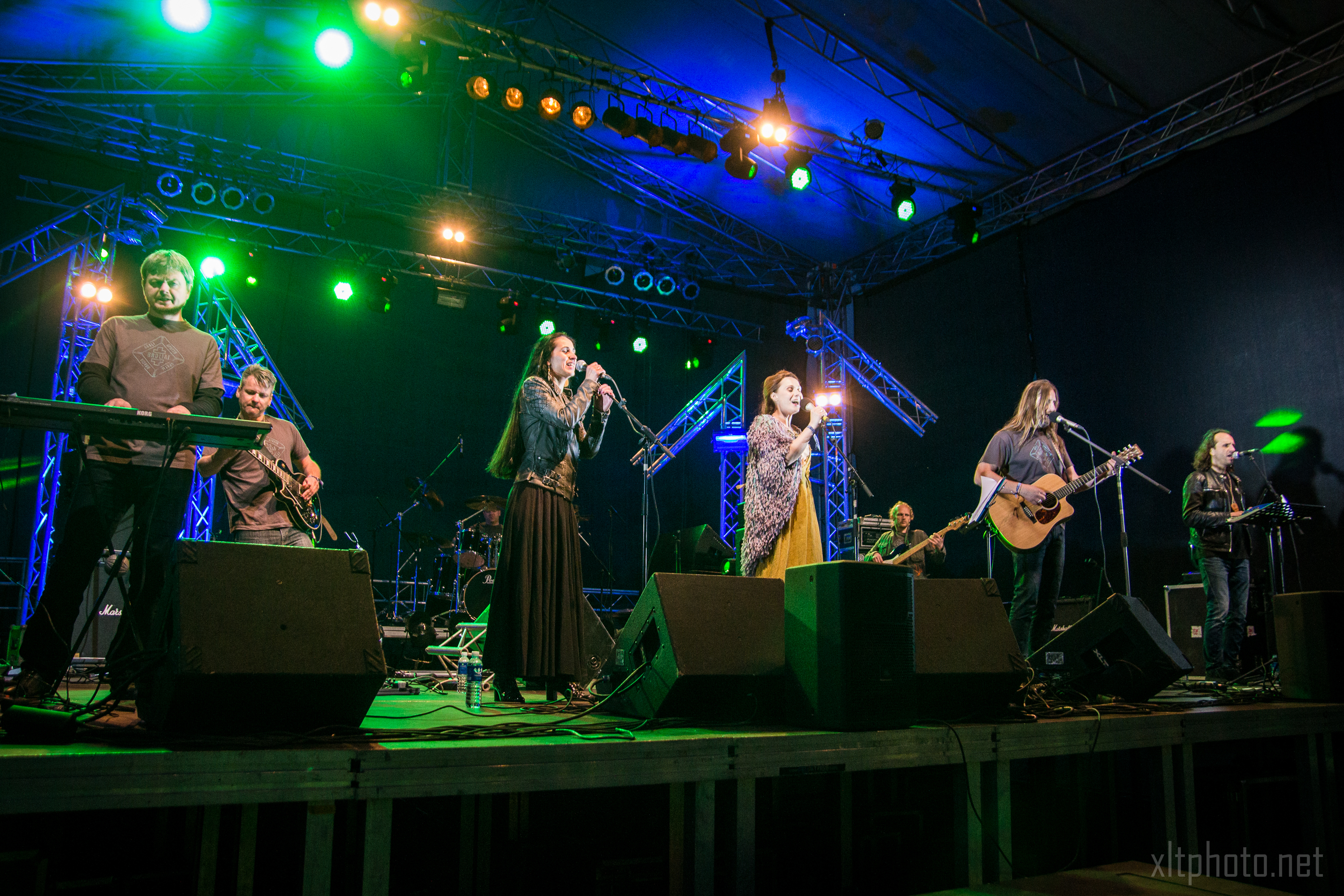 A band performs on an illuminated stage at Kilkim Žaibu 2015, with green, blue, and yellow lights. Six musicians—two vocalists, a guitarist, a bassist, a drummer, and a keyboardist—shine against the metal trusses and dark backdrop at this vibrant music festival.
