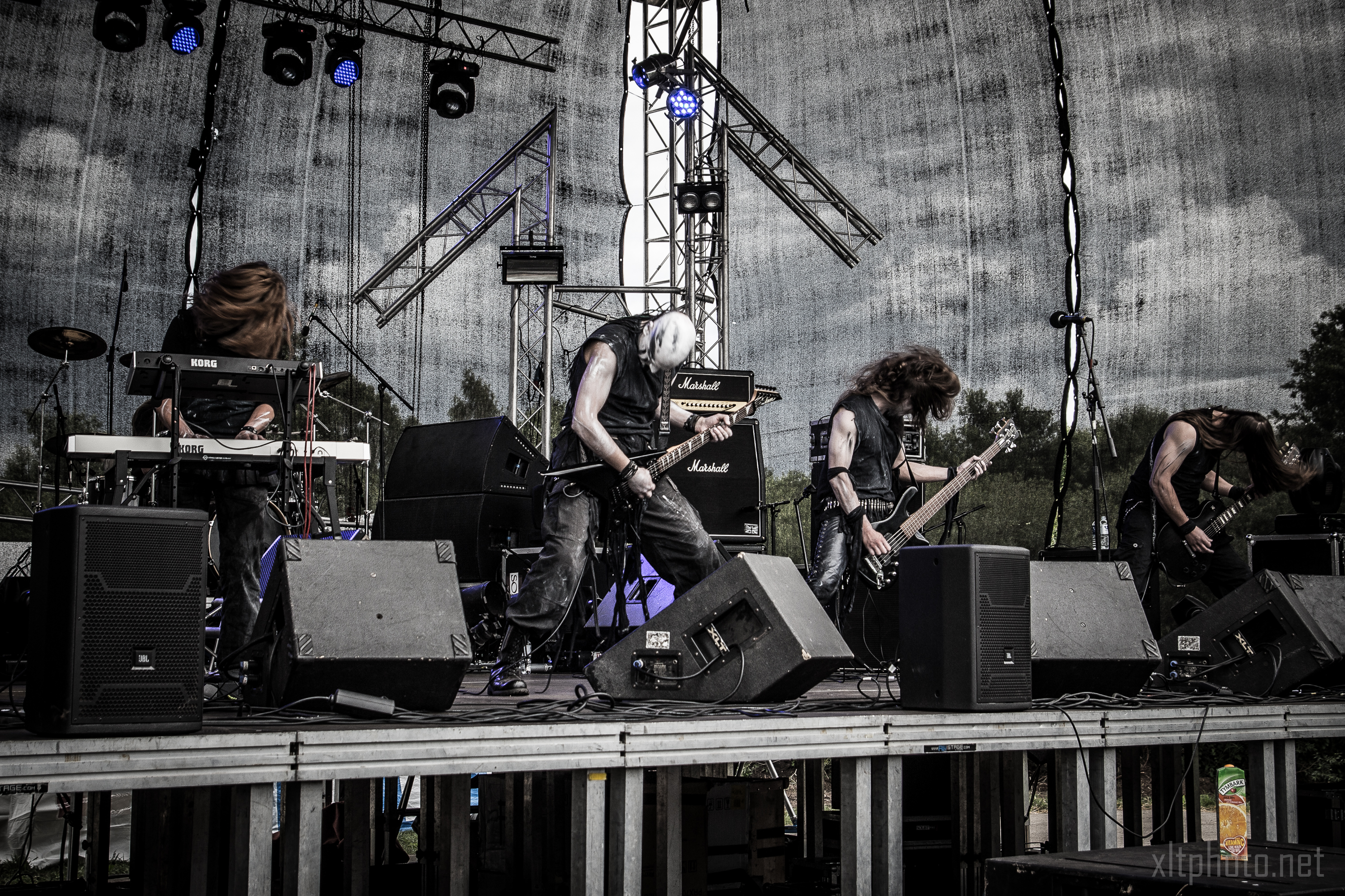 At the 2015 Kilkim Žaibu festival, a heavy metal band performs energetically onstage. Members include a keyboardist, two guitarists, and a drummer in the background. Amplifiers and speakers surround the stage. Intense lighting and a dark backdrop add to the moody, powerful atmosphere of the outdoor concert.