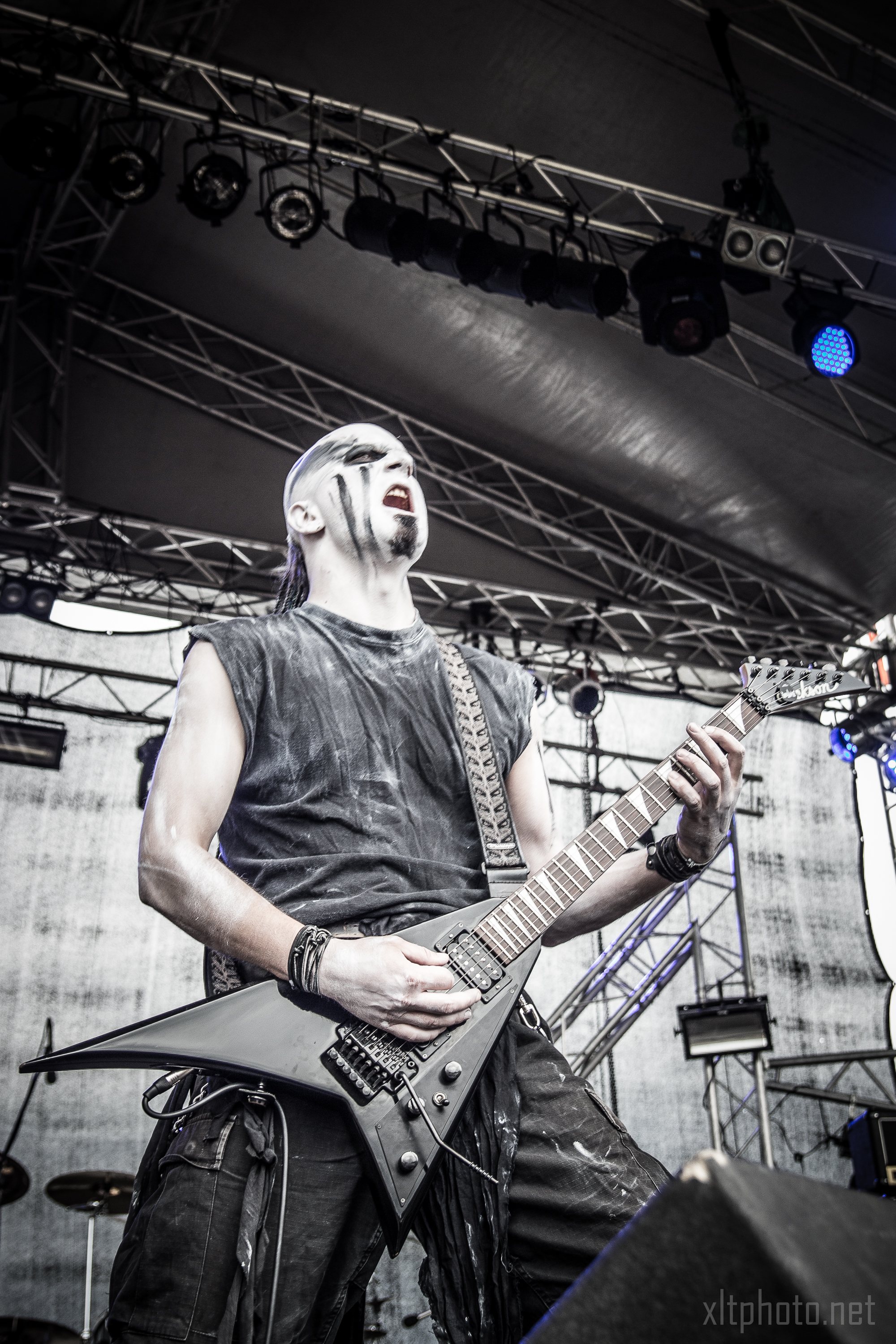 A musician passionately performs on stage at the 2015 Kilkim Žaibu event, playing an electric guitar. He wears black clothing and face paint that gives him a dramatic look. The background displays stage lighting and equipment, enhancing the energetic atmosphere of the live concert.