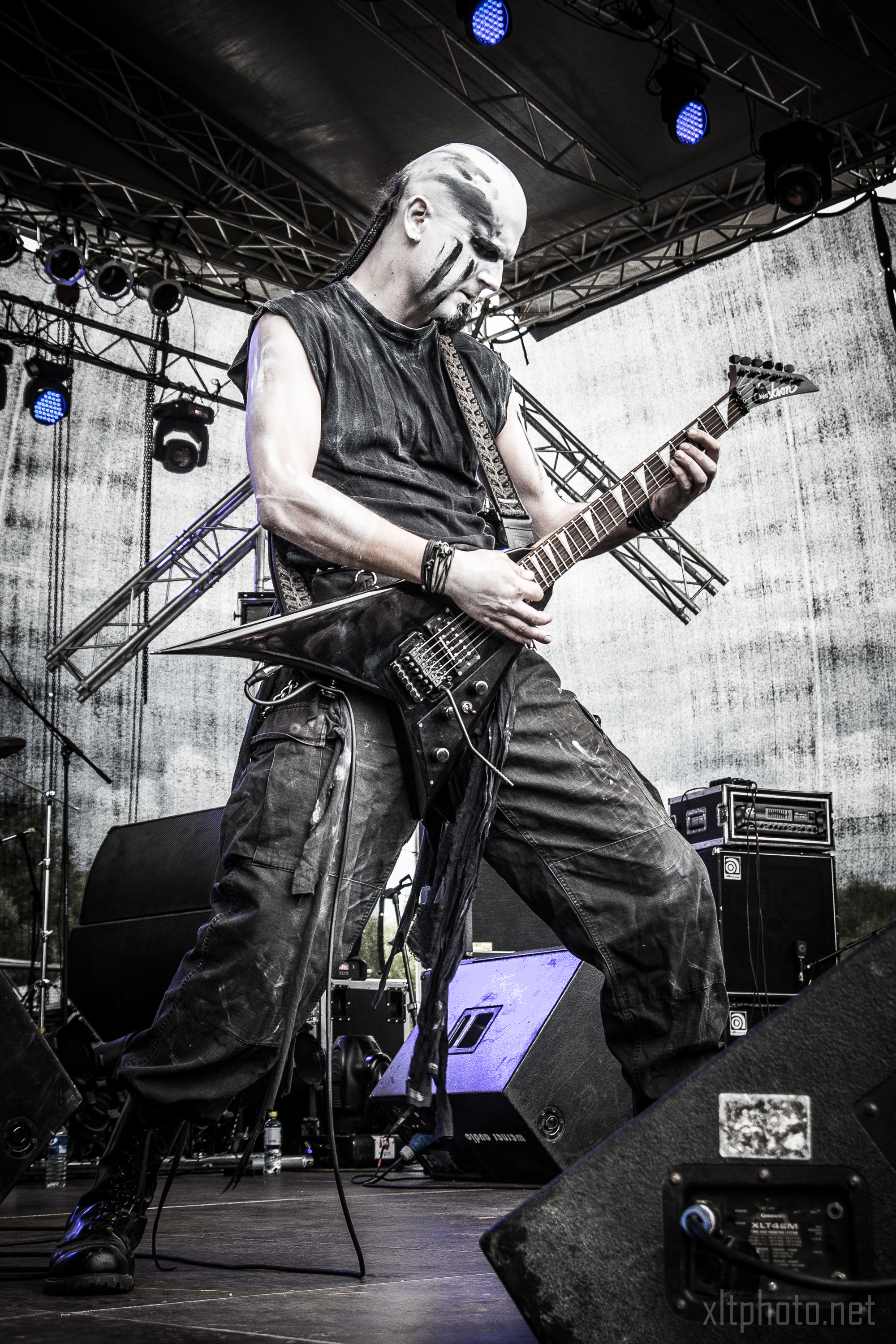 A musician with face paint, wearing dark, rugged attire, plays an electric guitar passionately on an outdoor stage at the 2015 Kilkim Žaibu festival. Bright stage lights illuminate him from above, and various stage equipment is visible. The background consists of metal scaffolding and a draped, semi-transparent tarp.