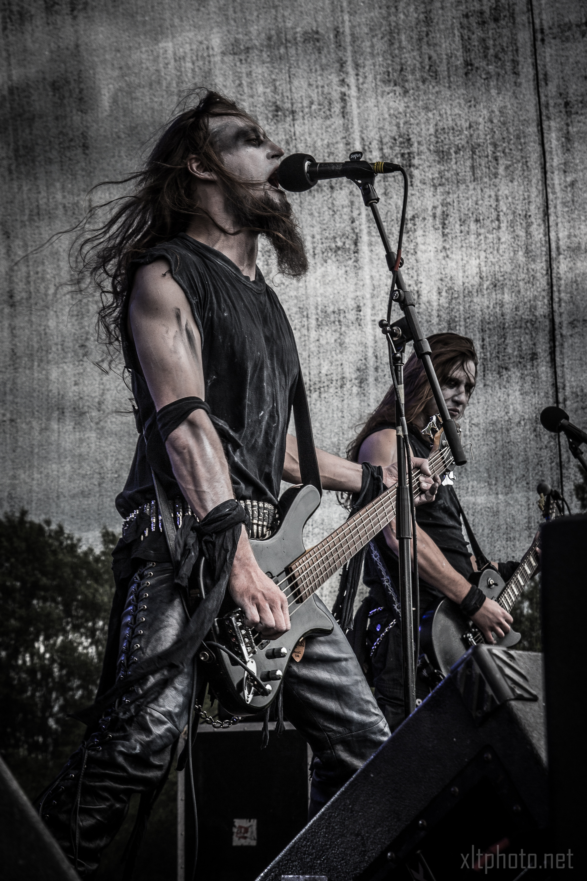 Two musicians perform energetically on stage at Kilkim Žaibu 2015, adorned in black metal attire with painted white and black faces. One sings into a microphone while playing a bass guitar, while the other strums an electric guitar. The backdrop is a gray, textured wall and trees are visible in the distance.