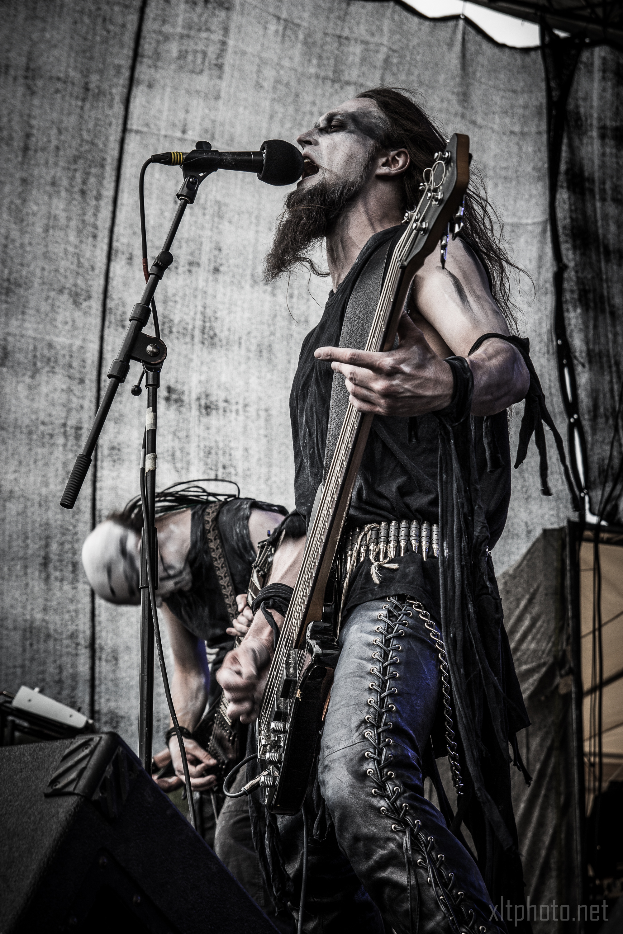 At the 2015 Kilkim Žaibu festival, a guitarist passionately performs on stage wearing black leather pants, a sleeveless shirt, wristbands, and face paint. The musician, with long hair and a beard, sings into a microphone while another band member plays guitar in the background. The setting is a live concert with a gritty, dark aesthetic.