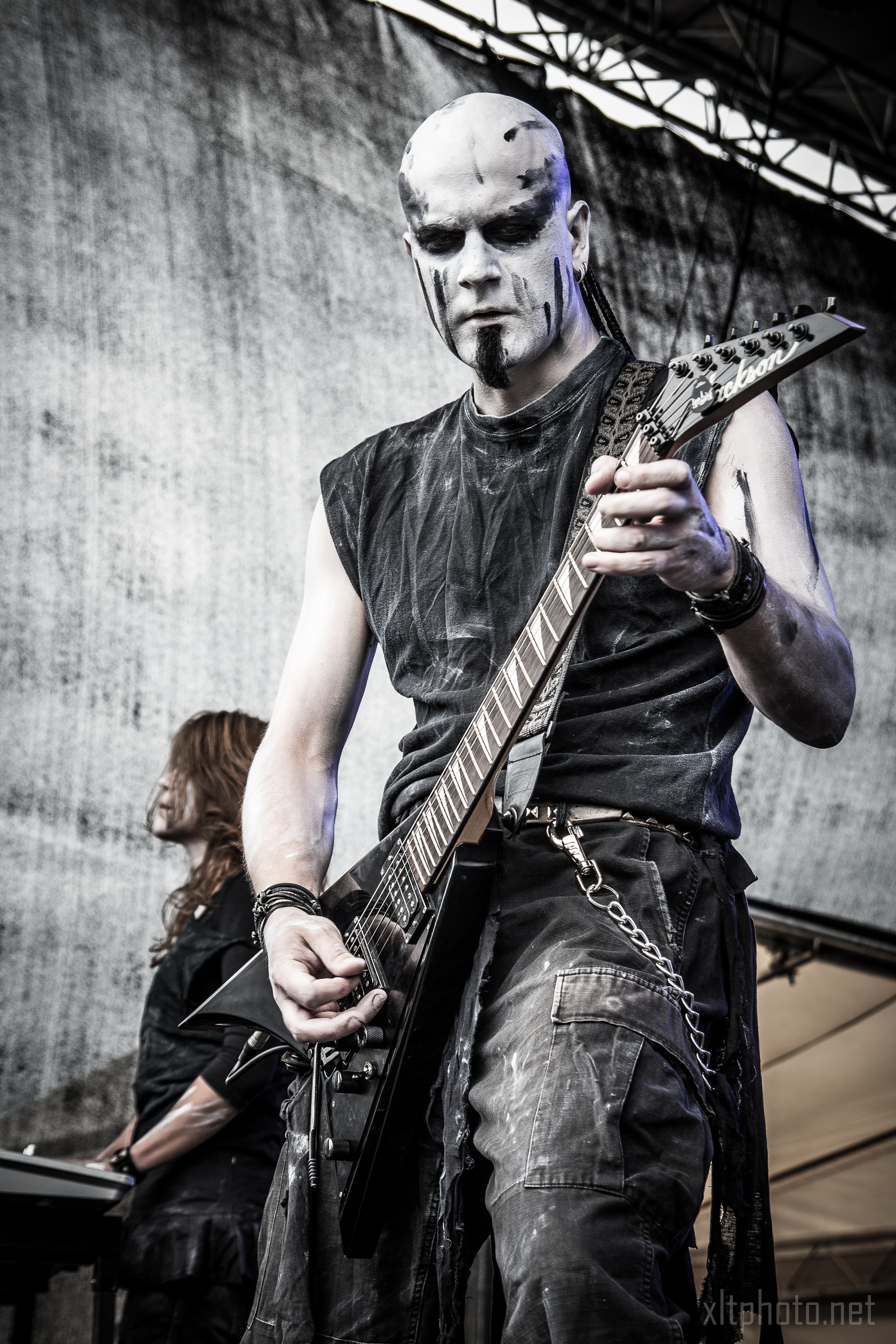 A guitarist with black-and-white face paint and black sleeveless attire performs on stage at the 2015 Kilkim Žaibu festival, gripping a black electric guitar. Background: a partially visible band member, drums, and stage equipment. The scene is set outdoors with a gray backdrop. A website watermark, "xlitephoto.net," is at the bottom right.