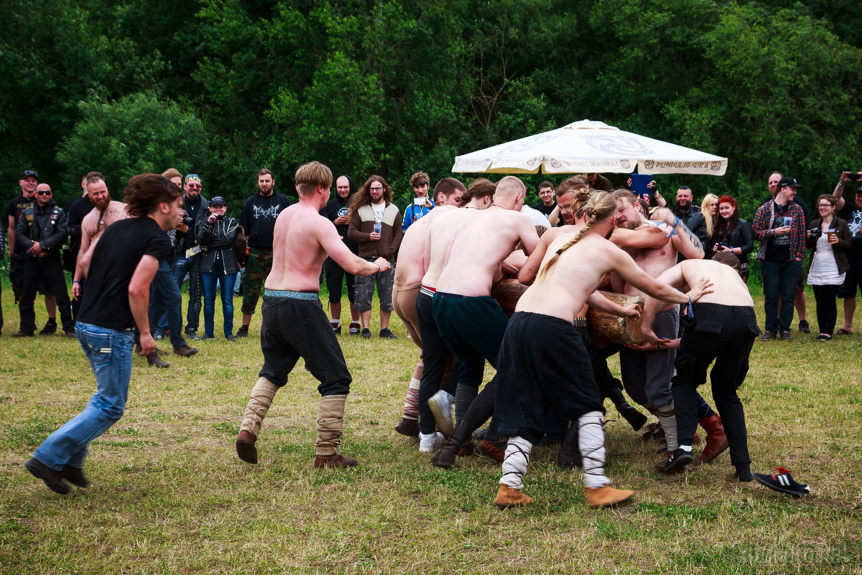 A group of shirtless people are participating in a wrestling match on grassy ground at the Kilkim Žaibu festival in 2015. Spectators, some dressed in casual attire while others wear themed costumes, stand around watching. A canopy umbrella is visible in the background amidst a green wooded area.