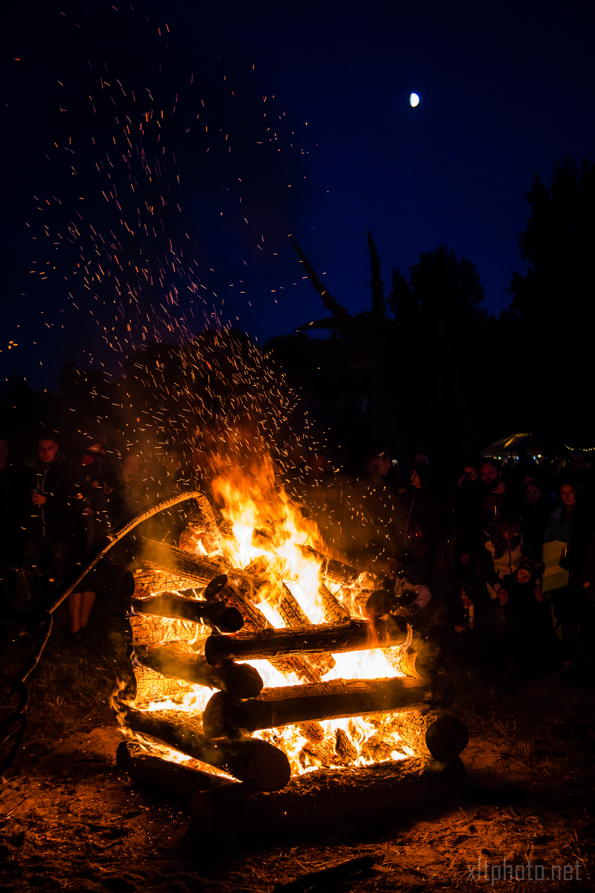 At the Kilkim Žaibu festival in 2015, a large bonfire burns brightly at night, with flames and sparks rising upwards. People are gathered around the fire, silhouetted against its light. The sky is dark blue, and a bright moon is visible above. Sparse trees and additional people can be seen in the background.