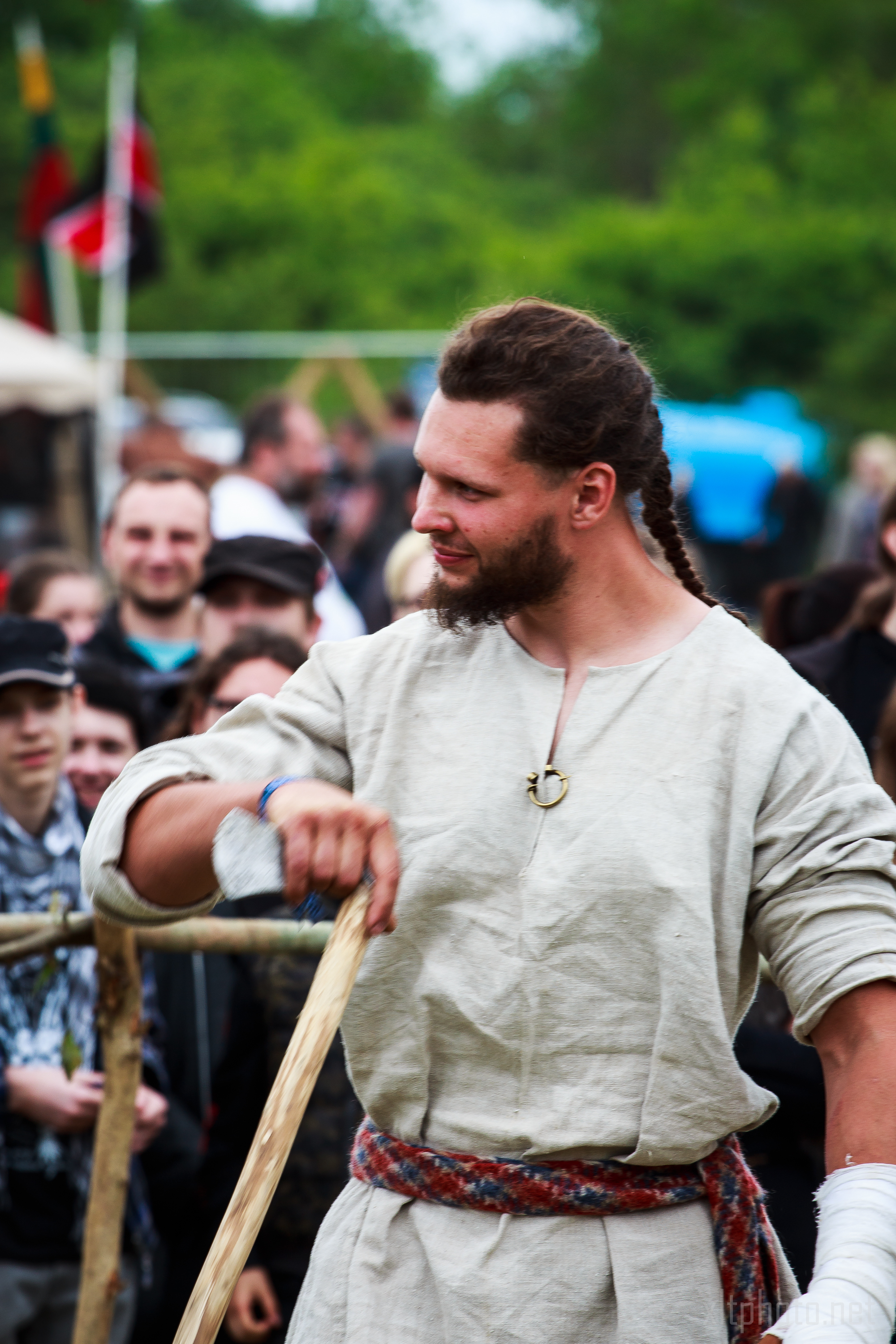 A man dressed in medieval attire stands outdoors at the 2015 Kilkim Žaibu festival, holding a wooden staff. He has long hair braided on one side, a beard, and is wearing a beige tunic with a decorative brooch and a woven belt. In the background, a crowd of people are watching, and there are trees and banners visible.