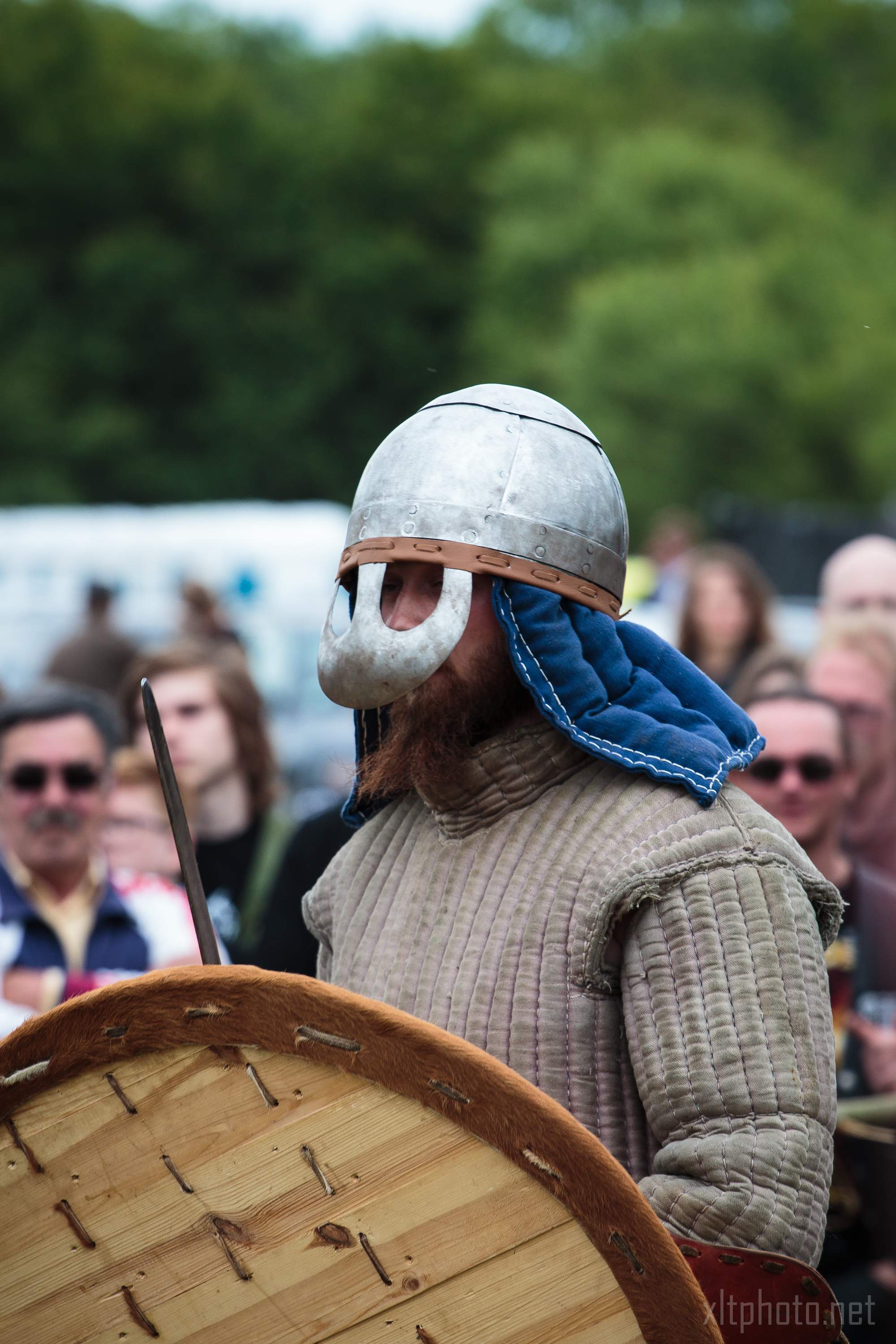 A person dressed in medieval armor stands in front of a blurred outdoor crowd at the 2015 Kilkim Žaibu festival. They wear a metal helmet with a protective face covering and a quilted garment with blue fabric lining at the neck, holding a large round wooden shield. Forested greenery is visible in the background.