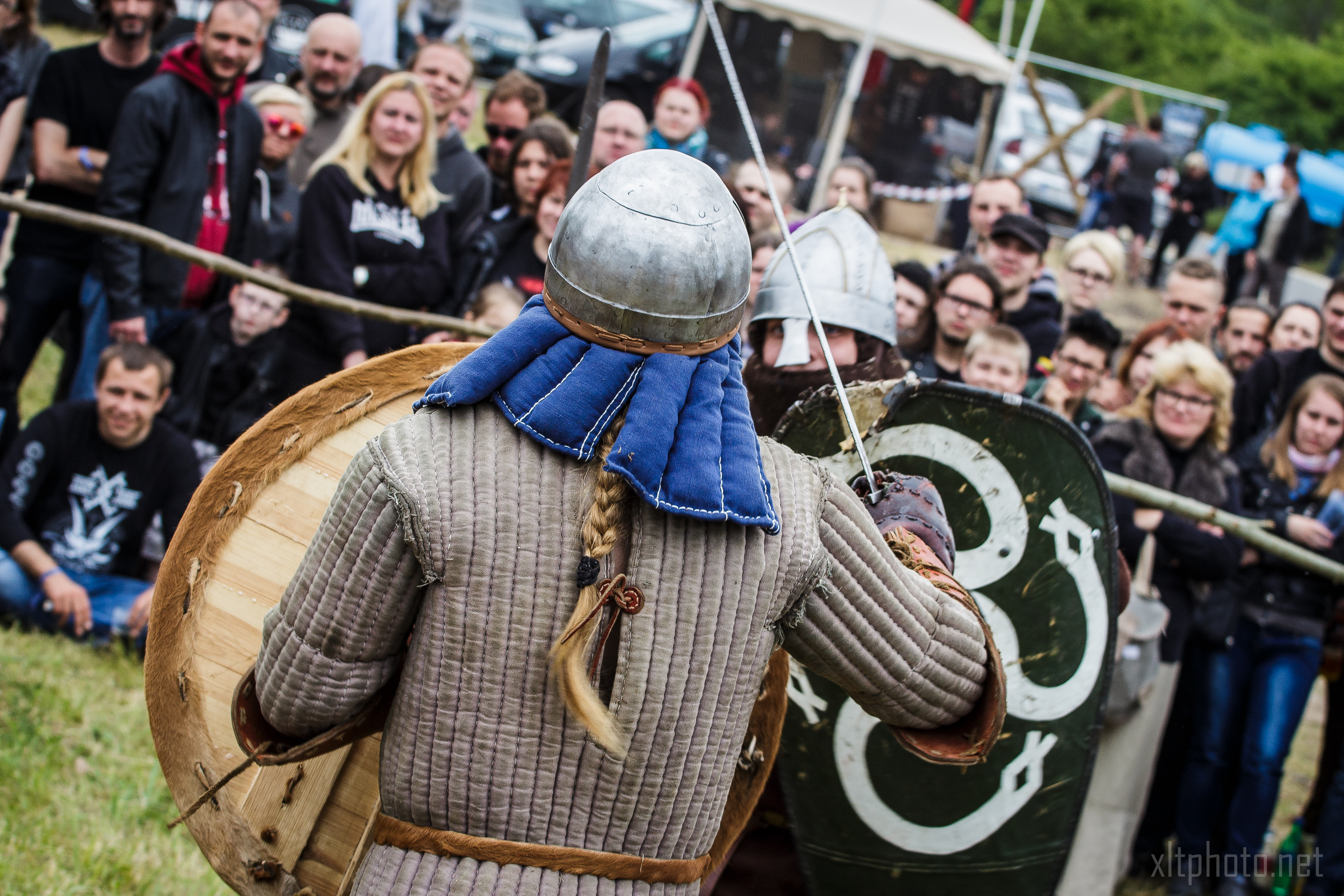 Two people in medieval armor, one with a long blonde braid, metal helmet, padded jacket, and shield, face each other in combat reenactment. A crowd at the 2015 Kilkim Žaibu festival watches intently from behind a rope barrier at the outdoor event. Tents and more spectators are visible in the background.