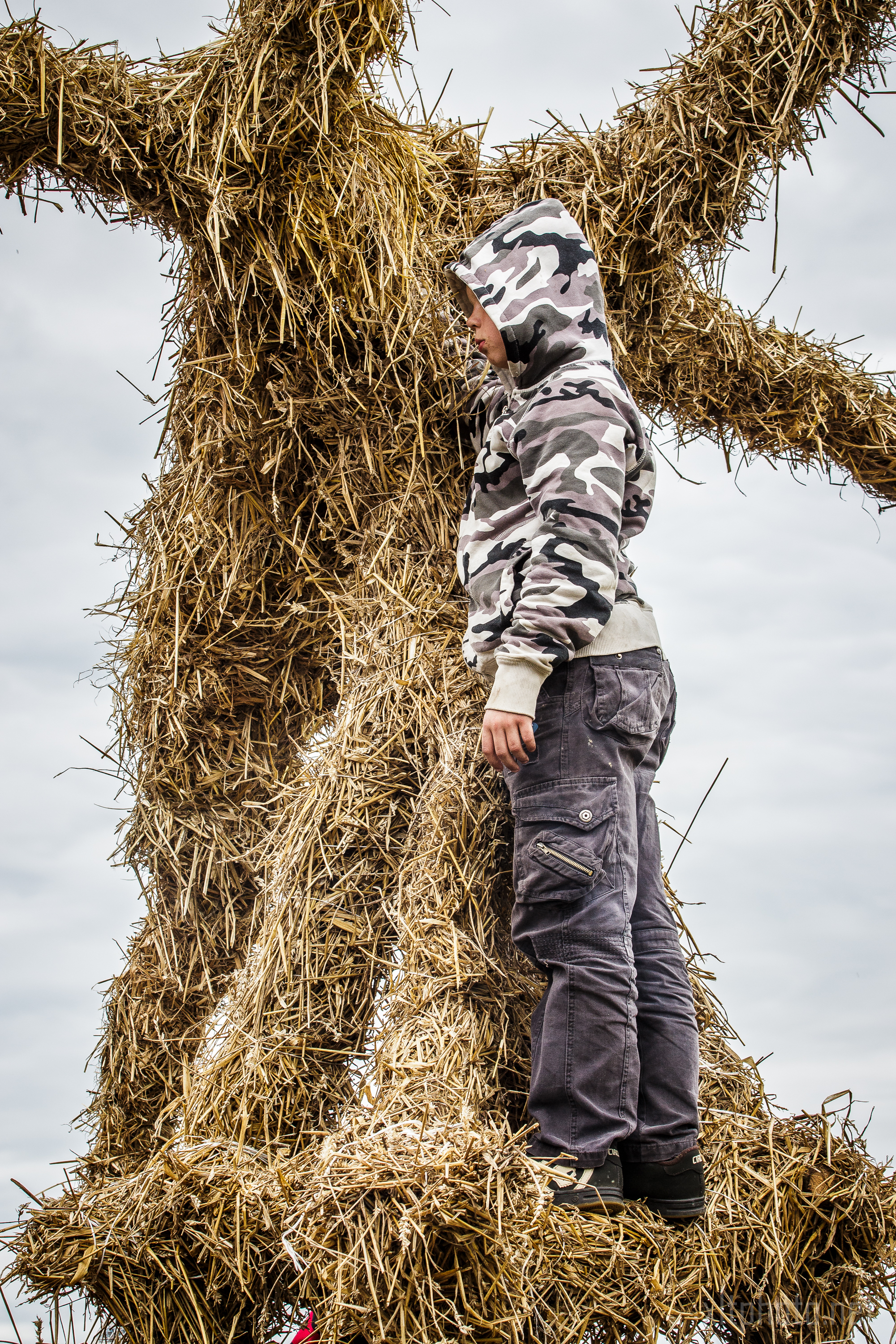 A person wearing a camouflage hoodie and dark cargo pants stands on a large, straw-covered structure at the 2015 Kilkim Žaibu festival. Their head is turned away, facing the textured straw surface, as an overcast sky looms in the background.