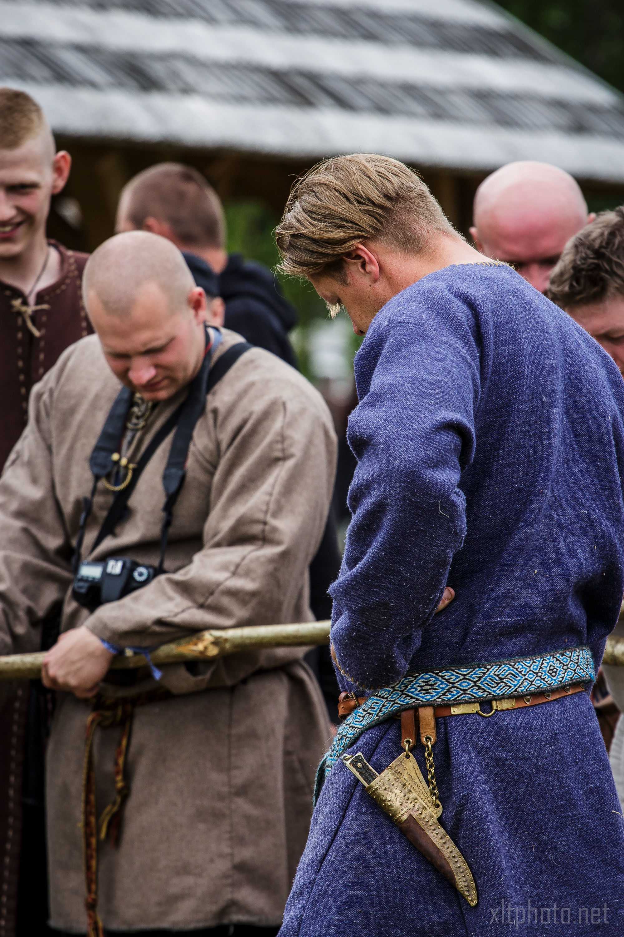 A group of men in medieval attire participates in an outdoor historical reenactment at the 2015 Kilkim Žaibu festival. The man in the foreground wears a blue tunic with a decorative belt, while the man to his left, in a brown tunic, holds a sword and has a camera around his neck. Other participants are partly visible in the background.