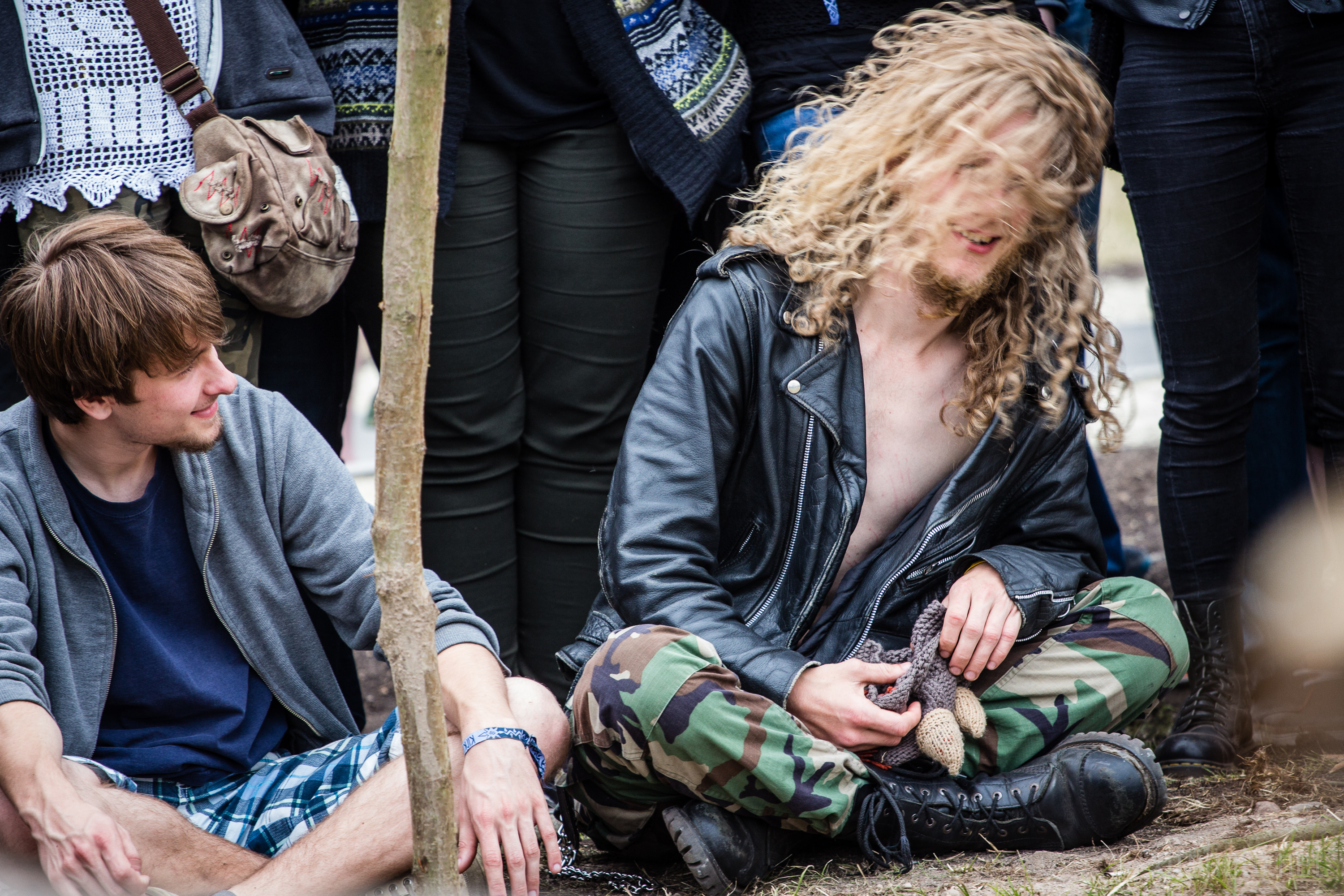 At the 2015 Kilkim Žaibu festival, a crowd is gathered outdoors. In the foreground, a person with long, curly blonde hair and a black leather jacket sits on the ground with legs crossed, wearing camouflage pants and black boots. To the left, another person wearing a gray hoodie and plaid shorts is sitting and smiling at them.