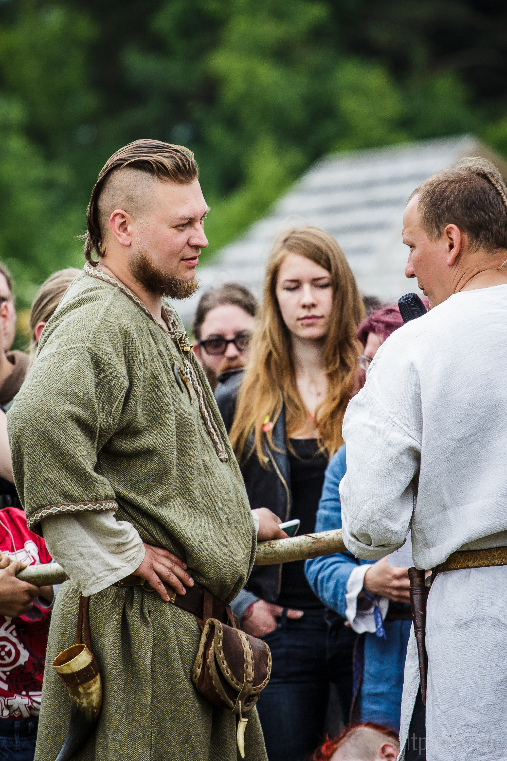 At the Kilkim Žaibu festival in 2015, a man with a short blonde mohawk, beard, and traditional green attire stands in a crowd, facing another man wearing a white robe and holding a microphone. Ornamental items hang from their belts. Onlookers, including a woman with long red hair, observe the scene against a blurry green background.