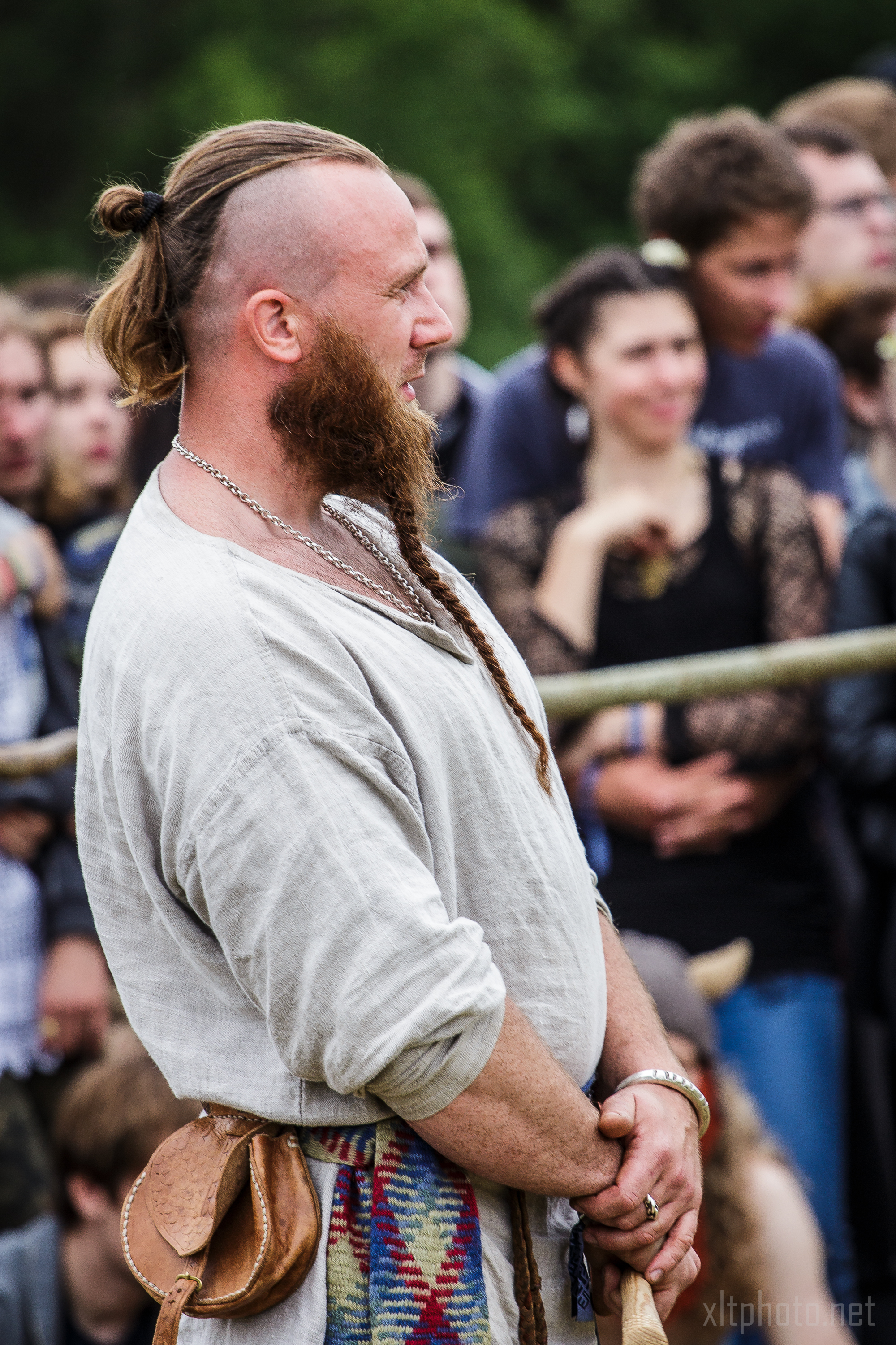 A man with a long beard and a partially shaved head, the remaining hair tied in a top knot, stands outdoors at the 2015 Kilkim Žaibu festival. He wears a loose, light-colored shirt and a woven belt with pouches. A crowd of people watches intently in the background.