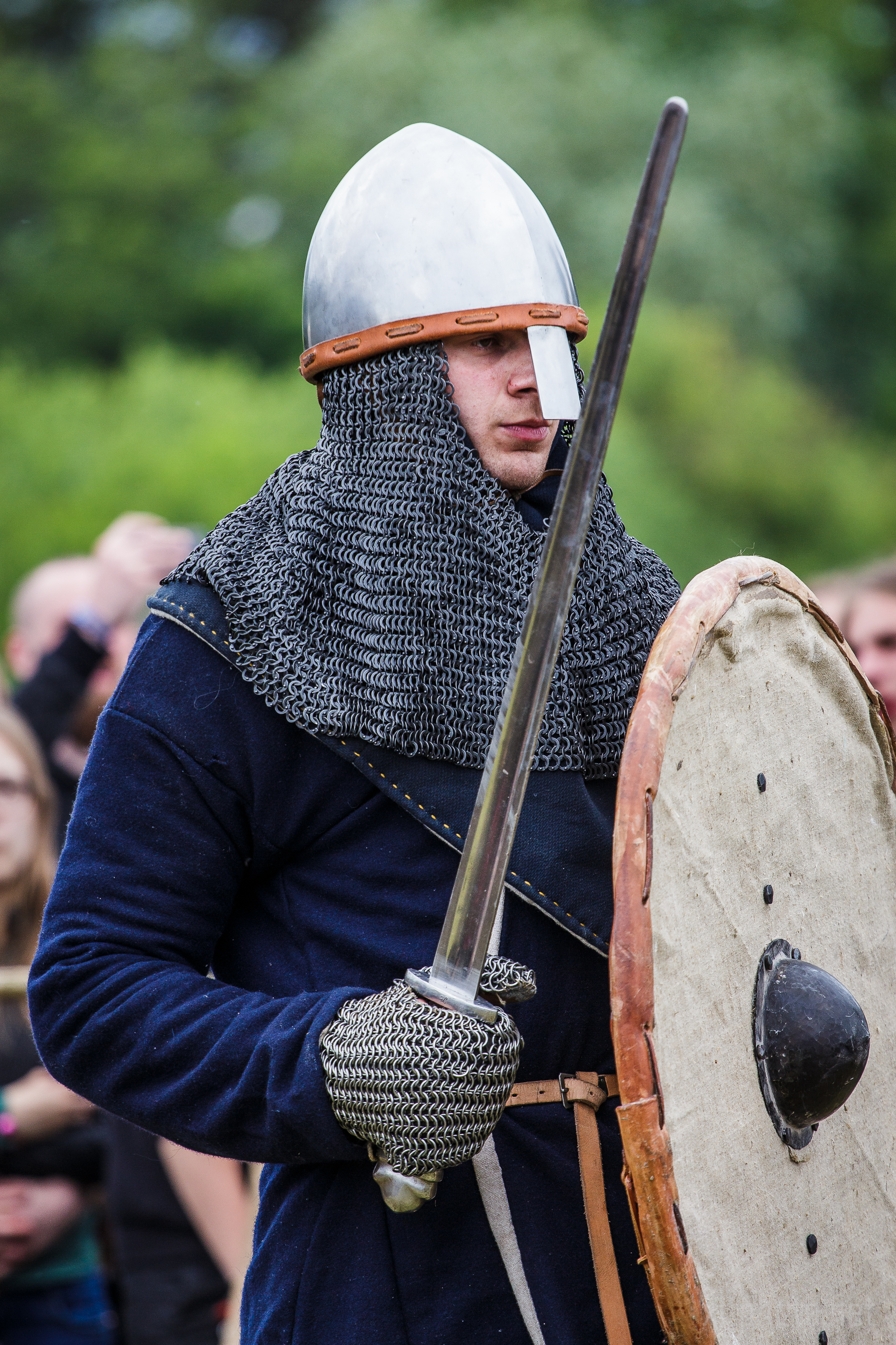 A person dressed in medieval armor holds a sword and a round shield. They wear a chainmail coif with a steel helmet and full medieval attire. The background shows an outdoor setting with blurred greenery and several people watching, reminiscent of the 2015 Kilkim Žaibu historical reenactment festival.
