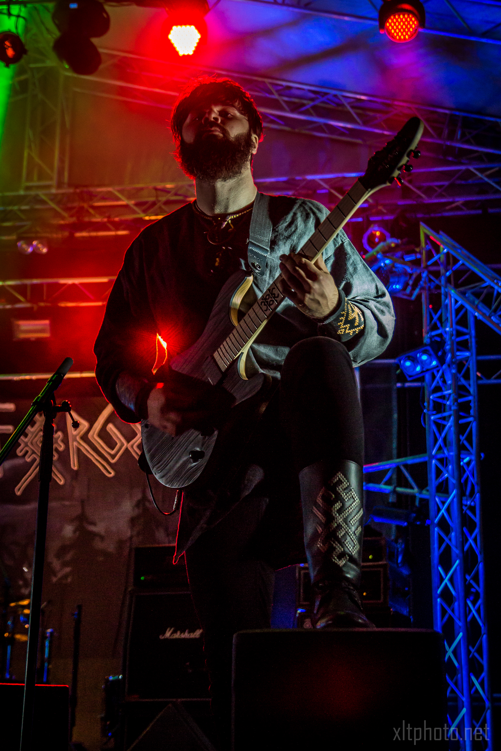 A guitarist with a beard and shoulder-length hair performs on stage with an electric guitar during the 2015 Kilkim Žaibu festival. He wears a dark shirt with embroidered sleeves, as stage lights in red, blue, and green illuminate the background. A blurred band logo and musical equipment are visible behind him.