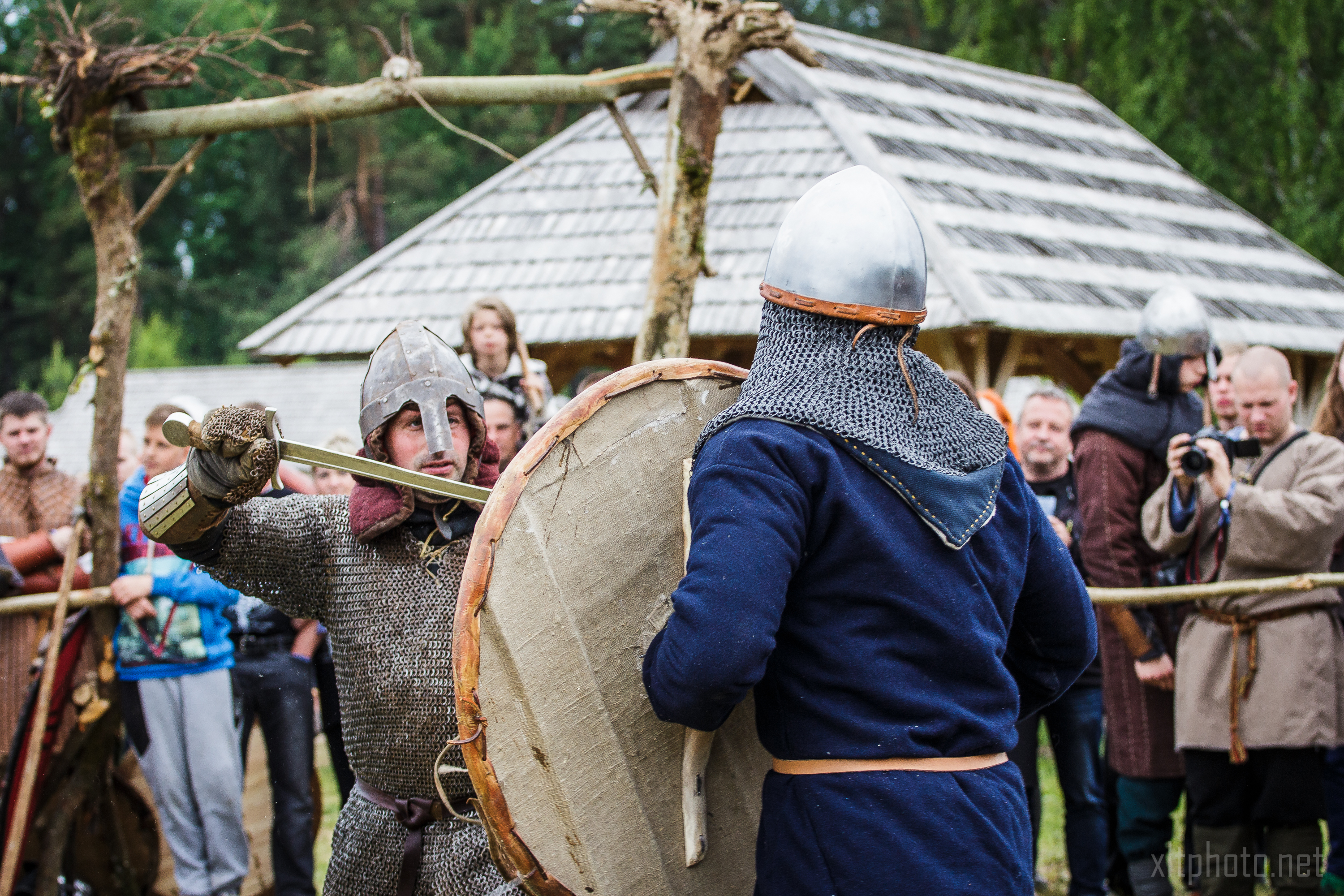 Two people in medieval combat attire engage in a mock sword fight during a reenactment event at Kilkim Žaibu 2015. They wear helmets and chainmail, and one holds a large round shield. Onlookers, some with cameras, observe the scene in front of rustic wooden structures set against a backdrop of trees.