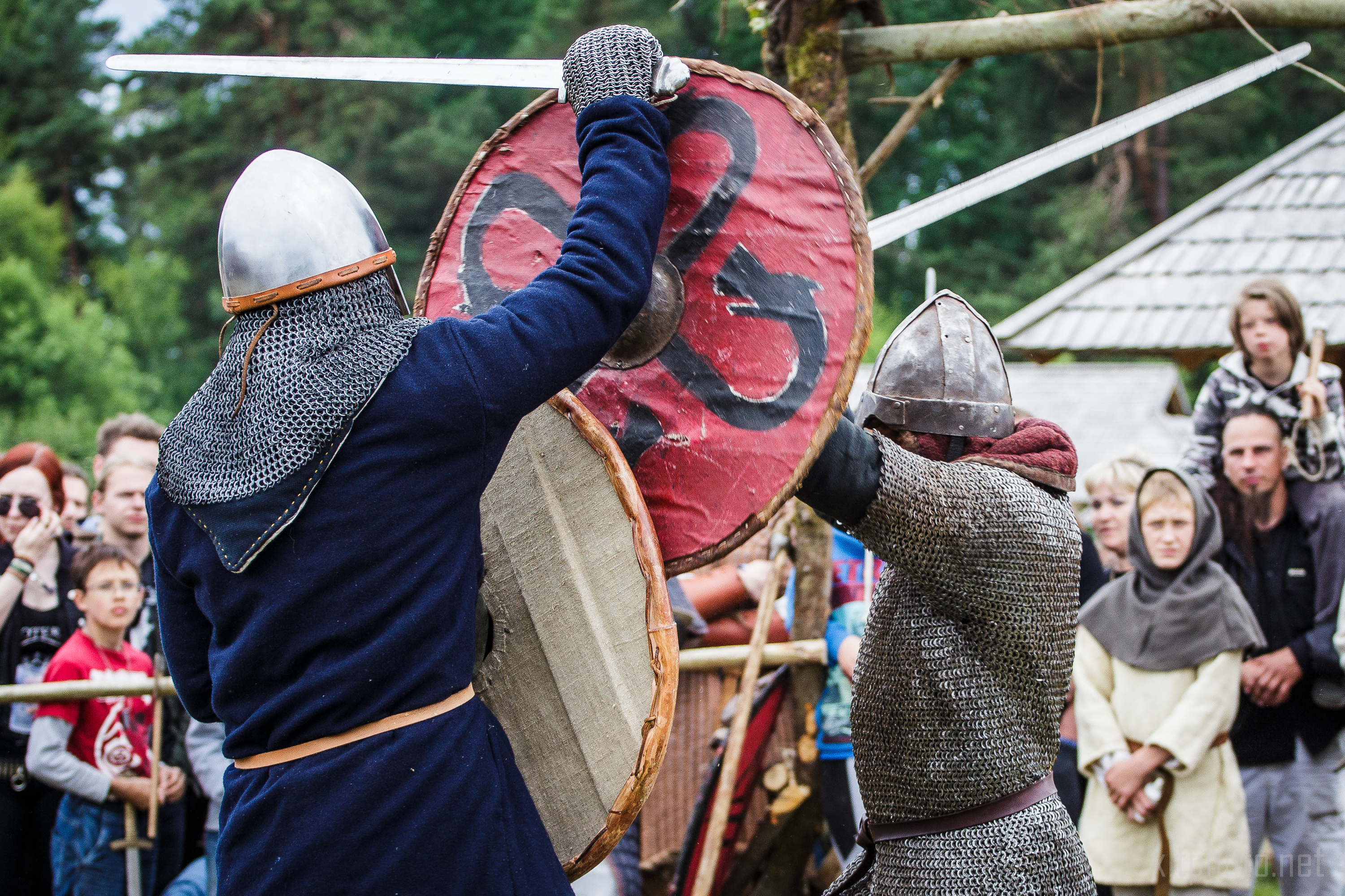 At the 2015 Kilkim Žaibu festival, two medieval reenactors, clad in chainmail and helmets, engage in combat. They wield combat shields and swords; one shield is painted with a prominent red design. Spectators, including adults and children, watch intently from the background, with trees and a rustic structure behind them.