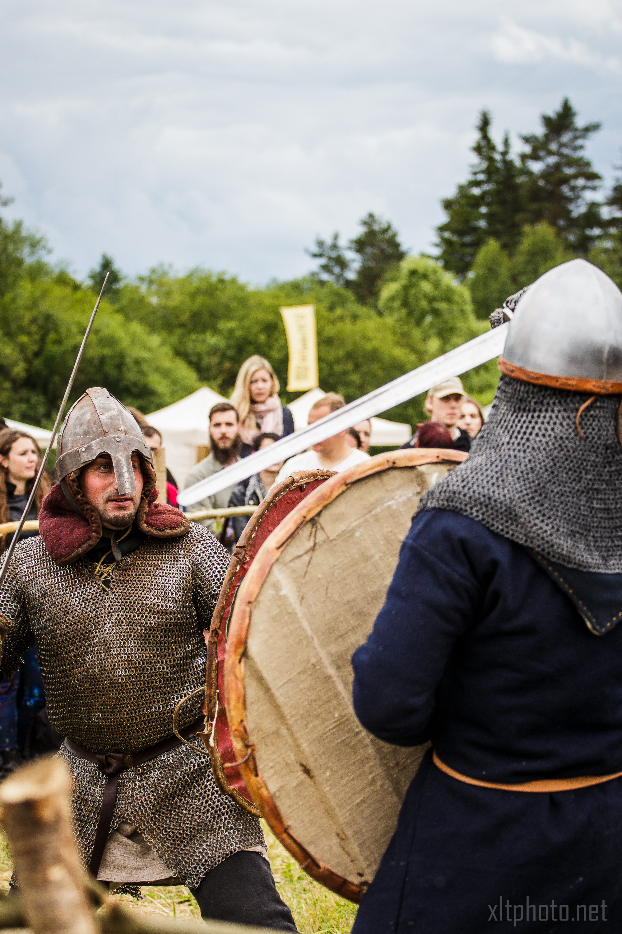 A medieval reenactment scene at the 2015 Kilkim Žaibu festival features two men in chainmail and helmets engaged in mock combat. One man holds a large round shield while the other thrusts a sword towards him. The background includes onlookers, tents, trees, and a cloudy sky, creating a lively historical atmosphere.