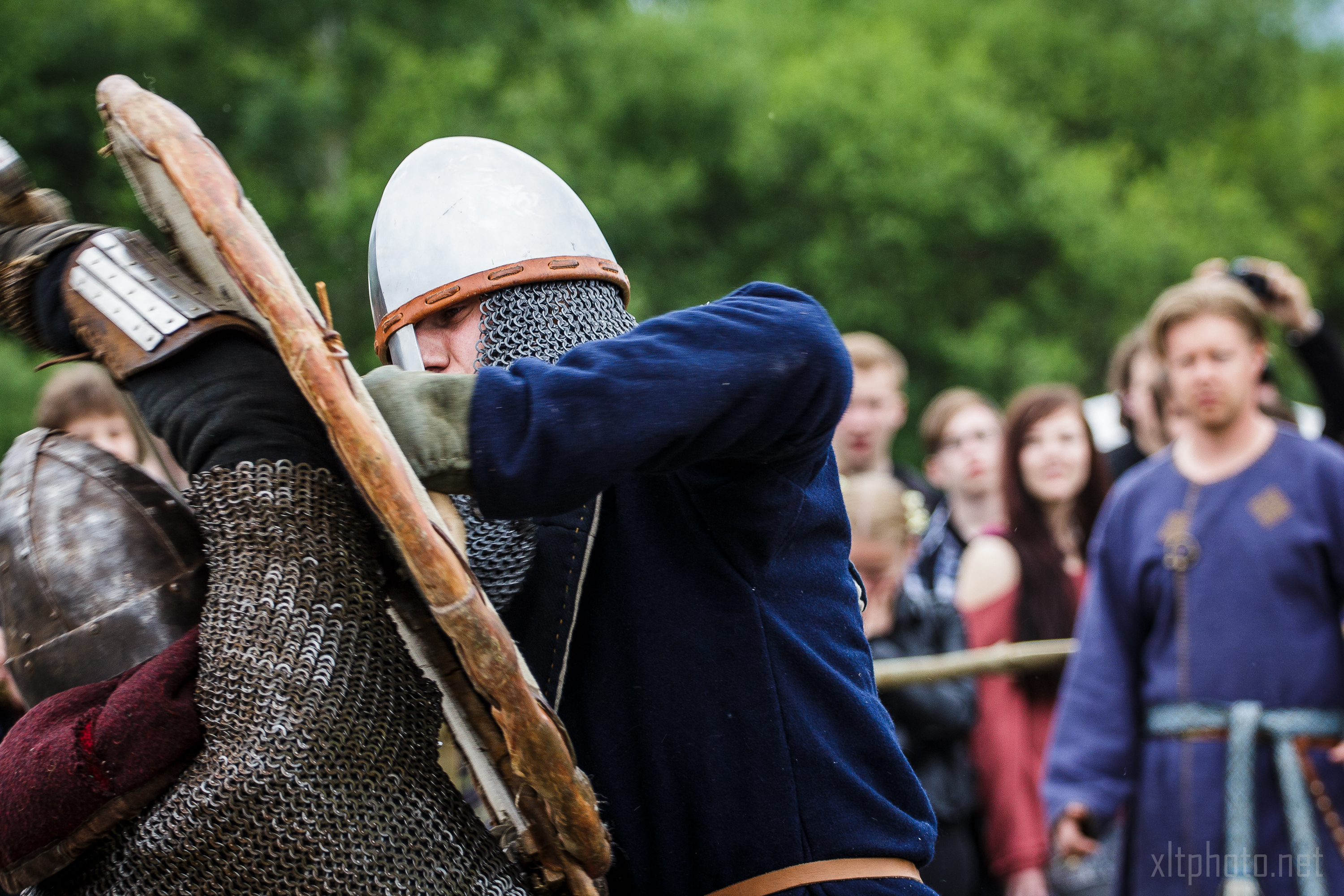 At the 2015 Kilkim Žaibu festival, two individuals clad in medieval armor, helmets, and chainmail engage in combat using wooden shields and swords. One is in a blue tunic, while the other is in red. A crowd of spectators, some taking pictures, watches against a backdrop of greenery.