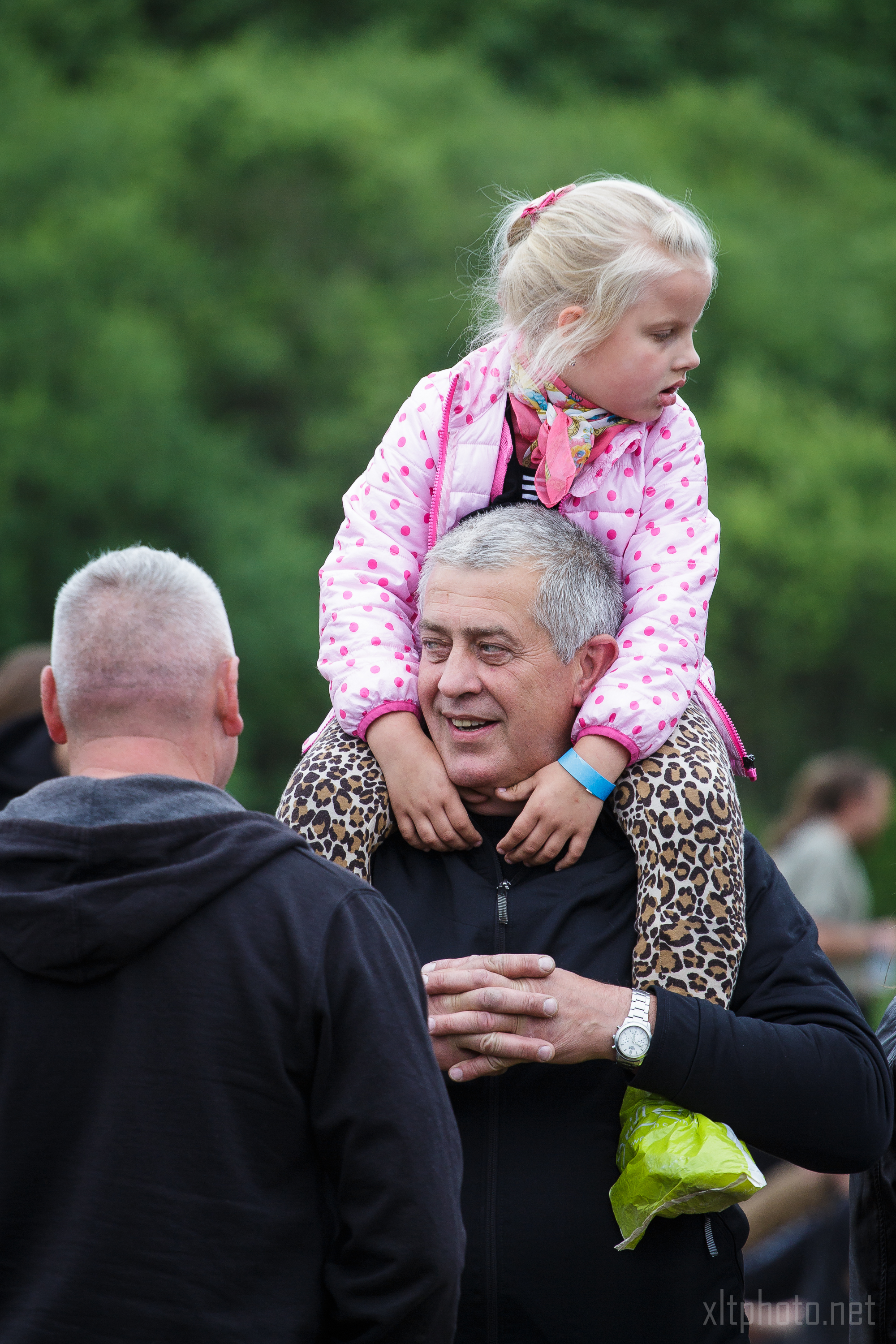 A gray-haired man smiles as he carries a young blonde girl on his shoulders. The girl is wearing a pink polka-dot jacket and leopard print pants. They are outdoors at the 2015 Kilkim Žaibu festival, with green trees visible in the background. Another man with short hair, facing away, is speaking to the man carrying the girl.