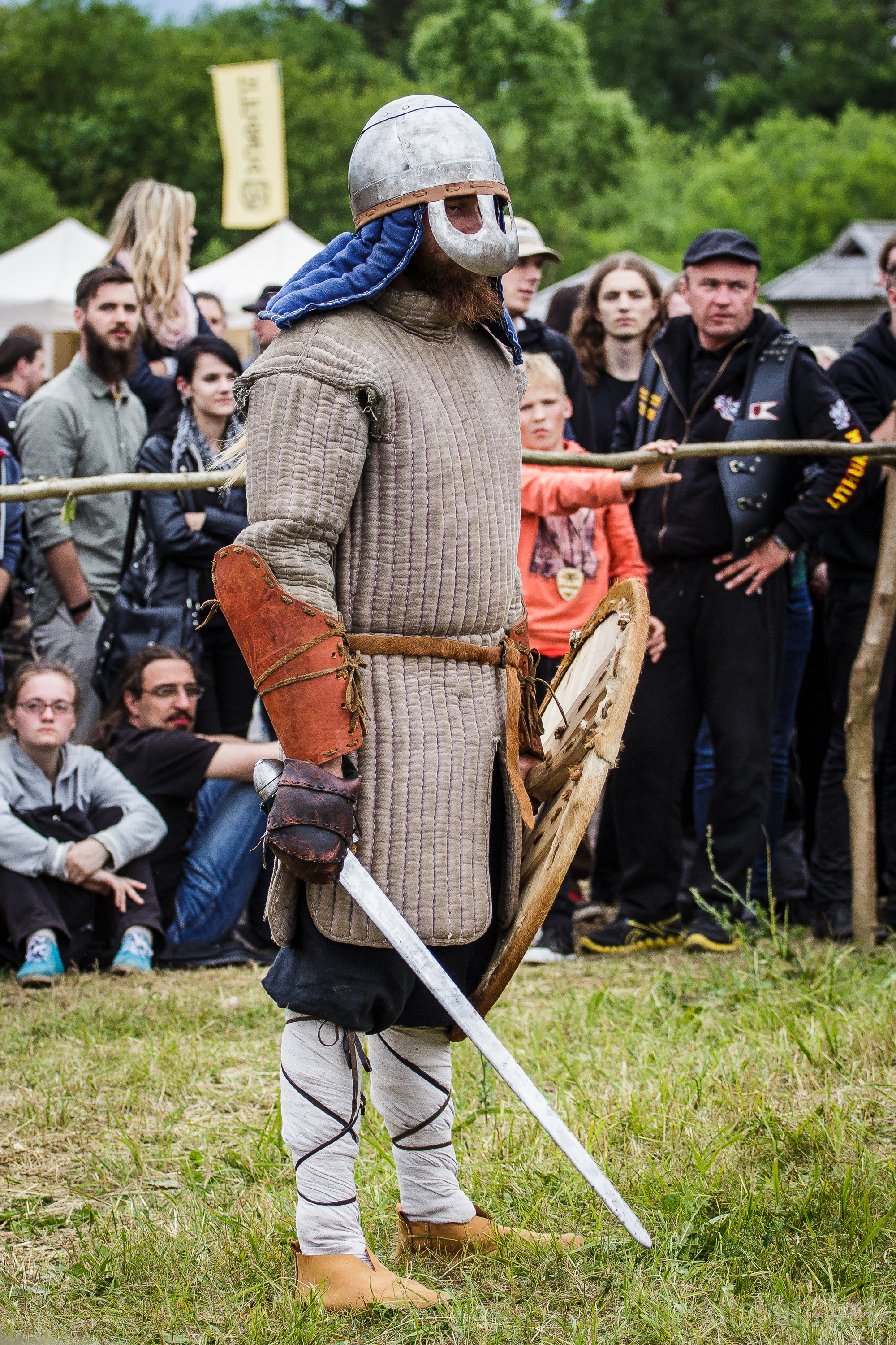 A person dressed in medieval armor, including a helmet with a face guard, padded tunic, and leather gauntlets, stands with a sword and wooden shield. Spectators at the 2015 Kilkim Žaibu festival watch attentively in the background, suggesting this scene is part of a historic reenactment.