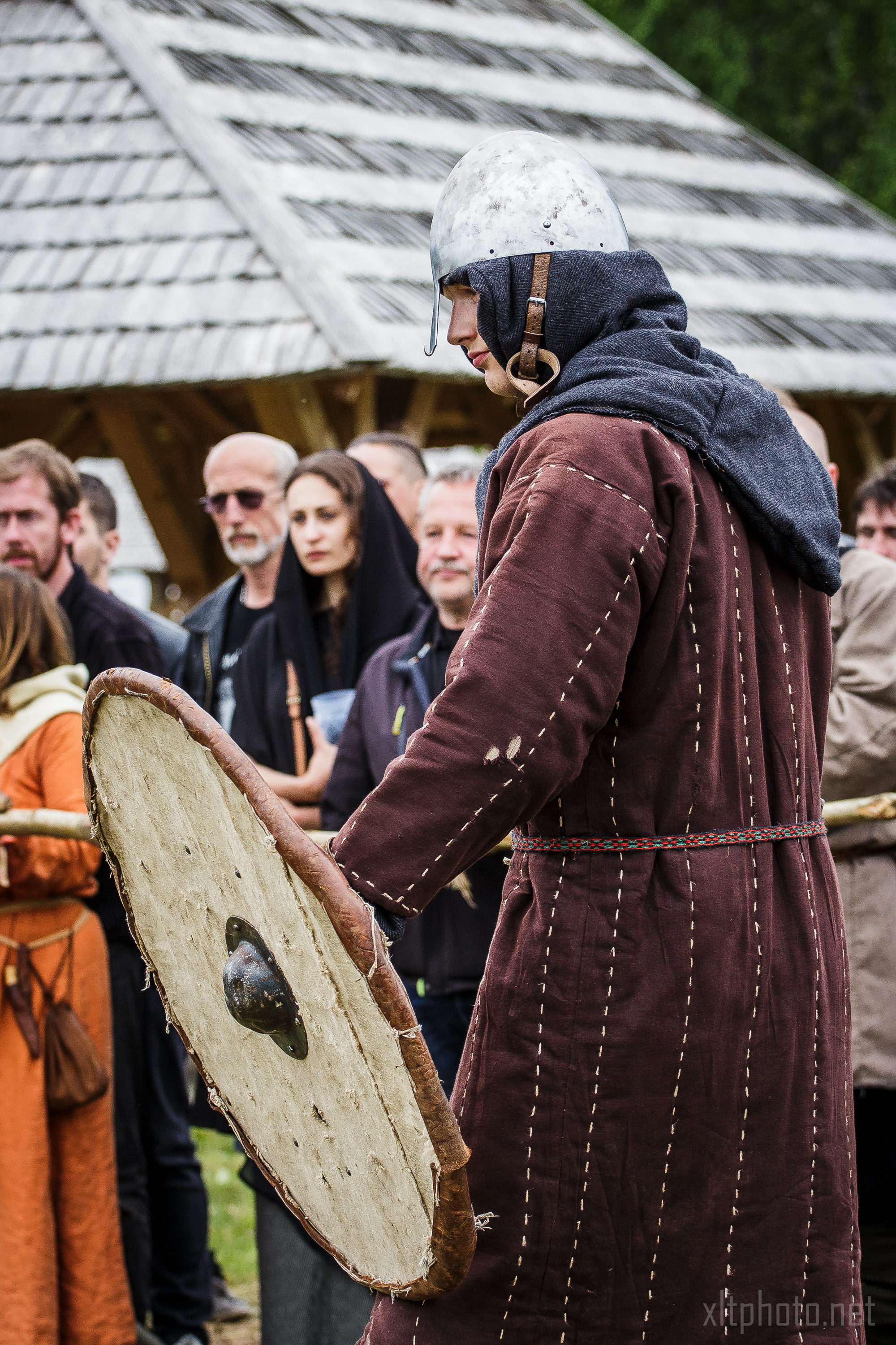 A person dressed in medieval clothing and a metal helmet stands holding a large, round shield. The individual is garbed in a brown tunic with a cloth hood and belt. In the background, attendees of Kilkim Žaibu 2015, clad in both period attire and modern clothes, gather near a wooden structure with a shingled roof.